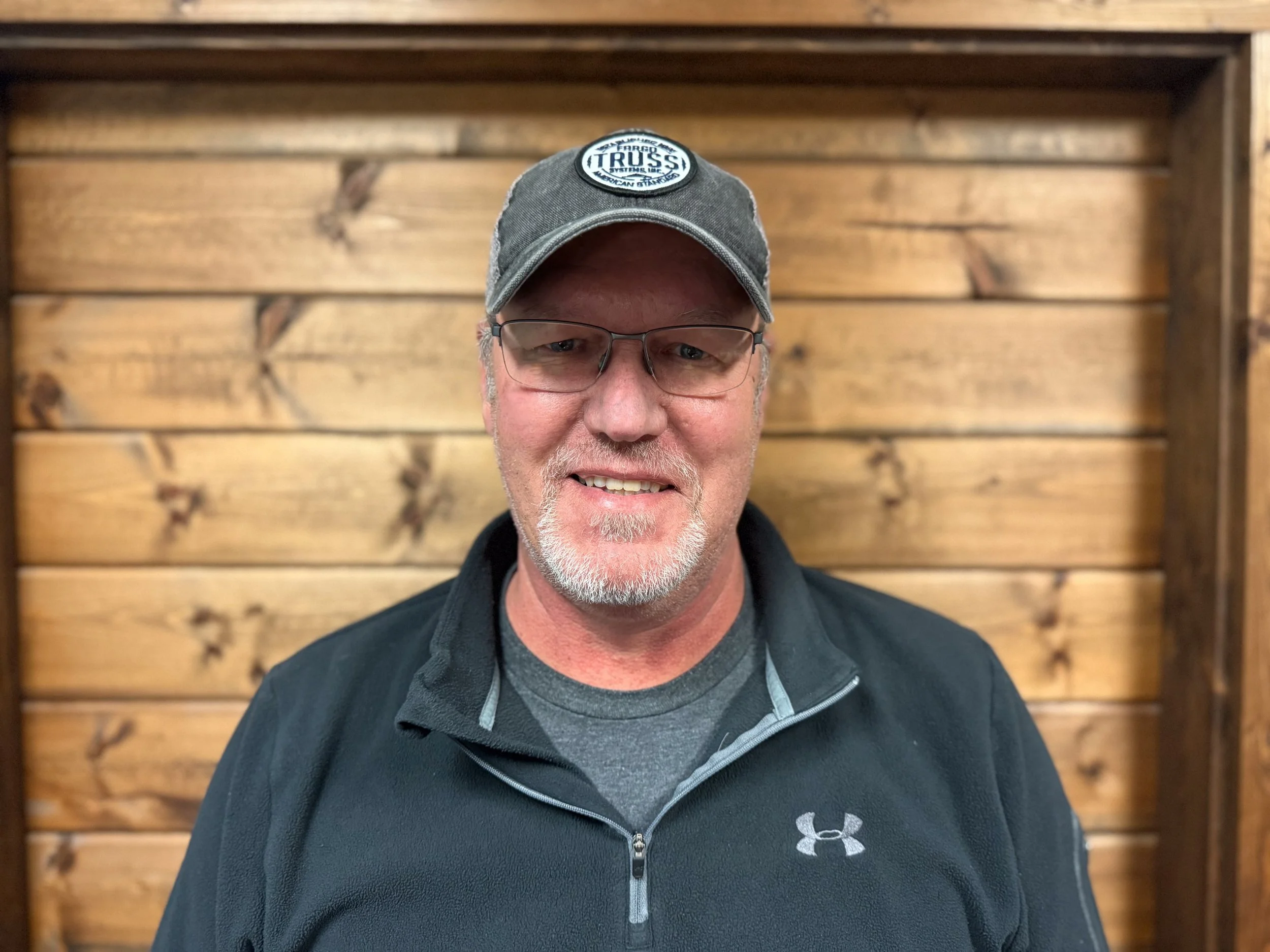 A smiling middle-aged man with glasses, a gray beard, and wearing a gray baseball cap with a patch and a black Under Armour zip jacket, standing in front of a wooden wall.