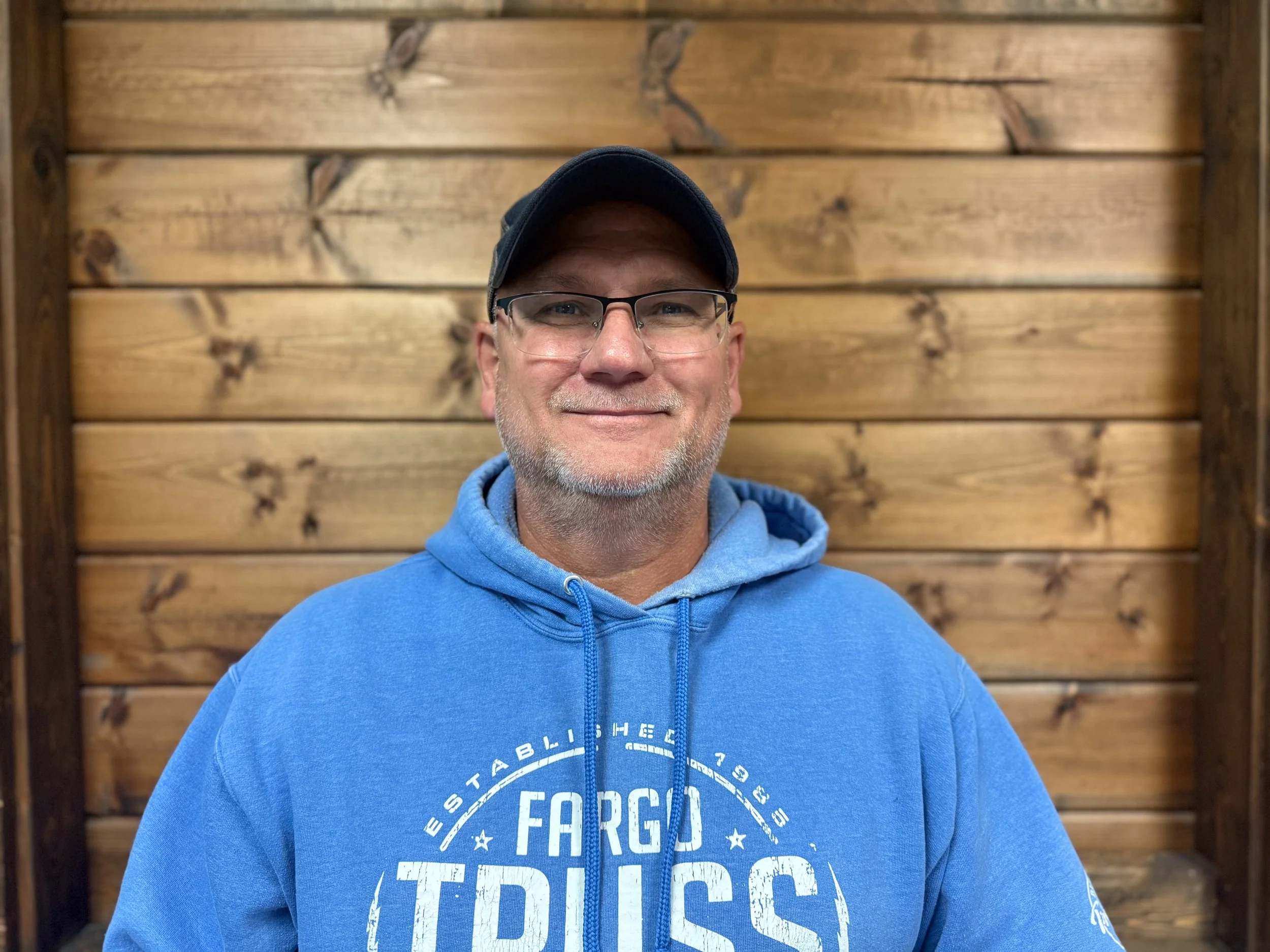A middle-aged man with glasses, a gray beard, wearing a black cap and a blue hoodie, standing in front of a wooden wall, smiling at the camera.