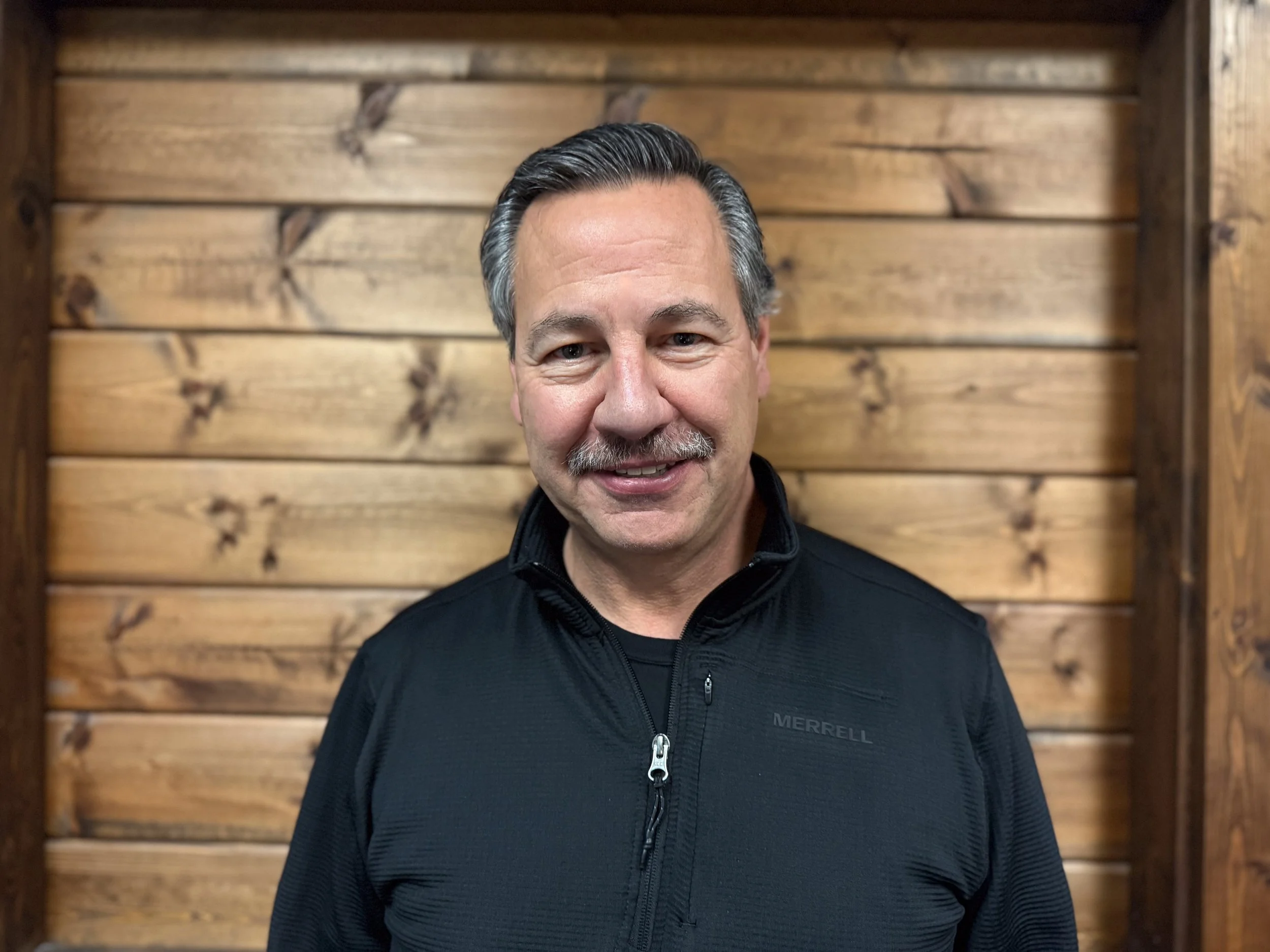 A middle-aged man with gray hair and a mustache, wearing a black zip-up jacket, standing in front of a wooden wall.