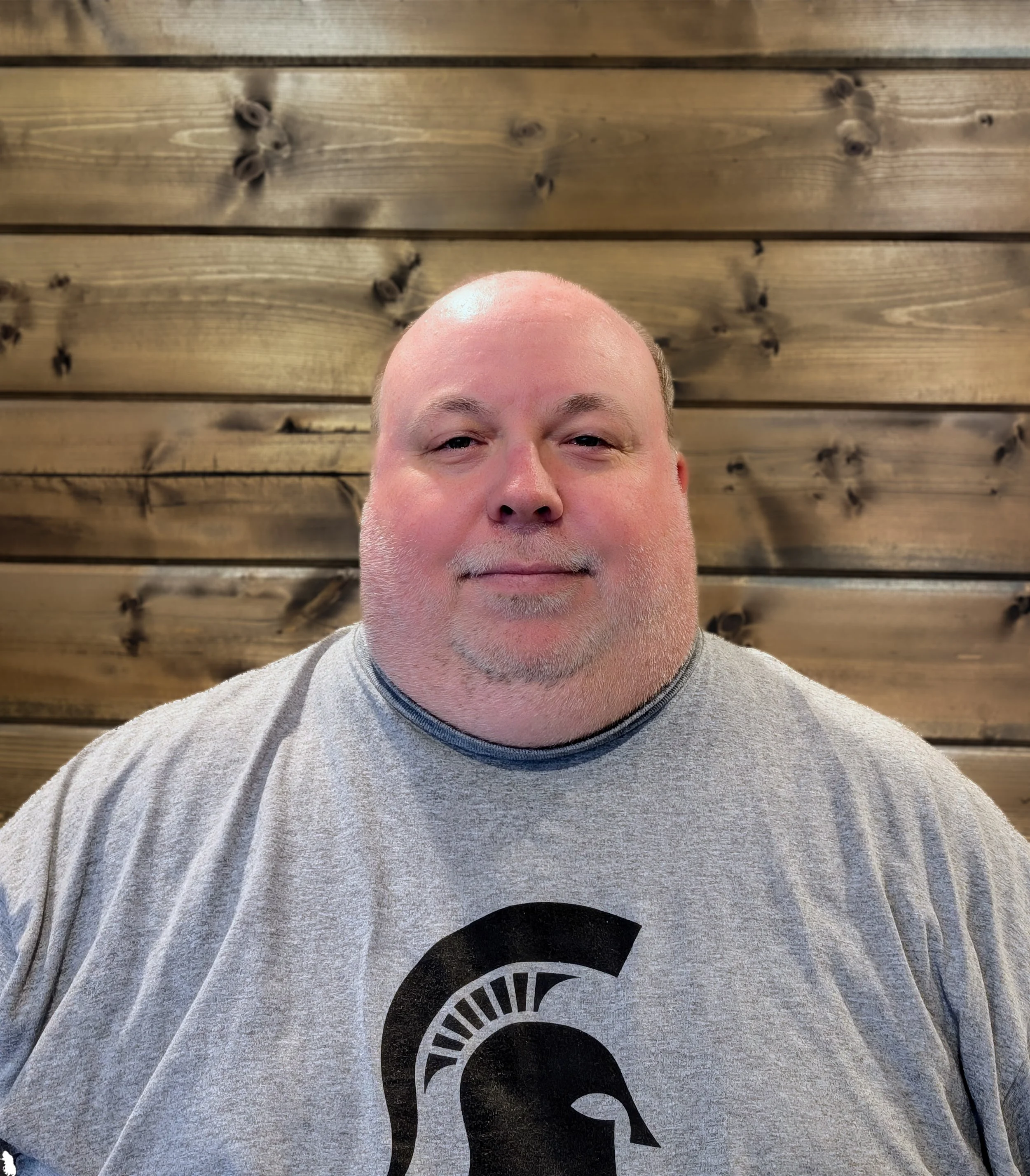 A man with a shaved head and a beard, wearing a gray T-shirt with a black Spartan helmet logo, standing in front of a wooden wall.