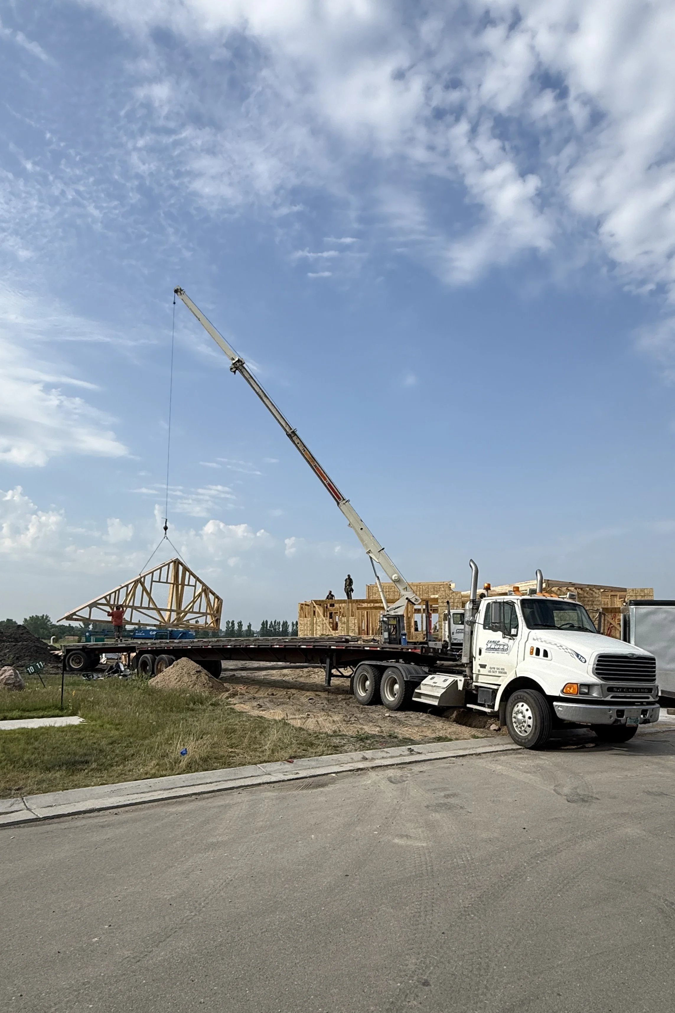 Construction site with workers and a crane lifting a wooden house frame, truck parked nearby, and ongoing building framework against partly cloudy sky.