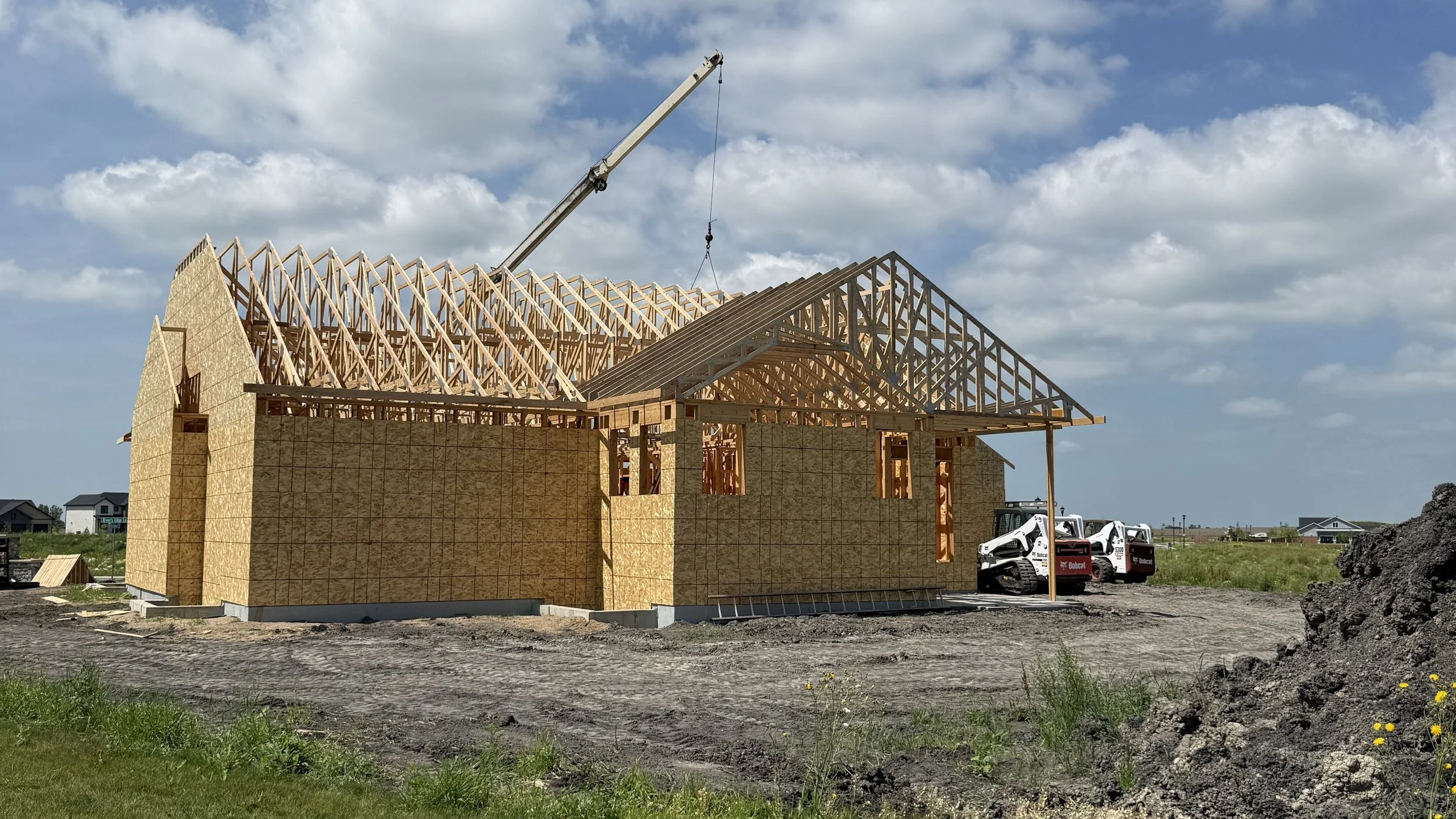 Construction site with a wooden house frame and roof trusses being assembled, a crane lifting materials, and construction equipment on a clear day.