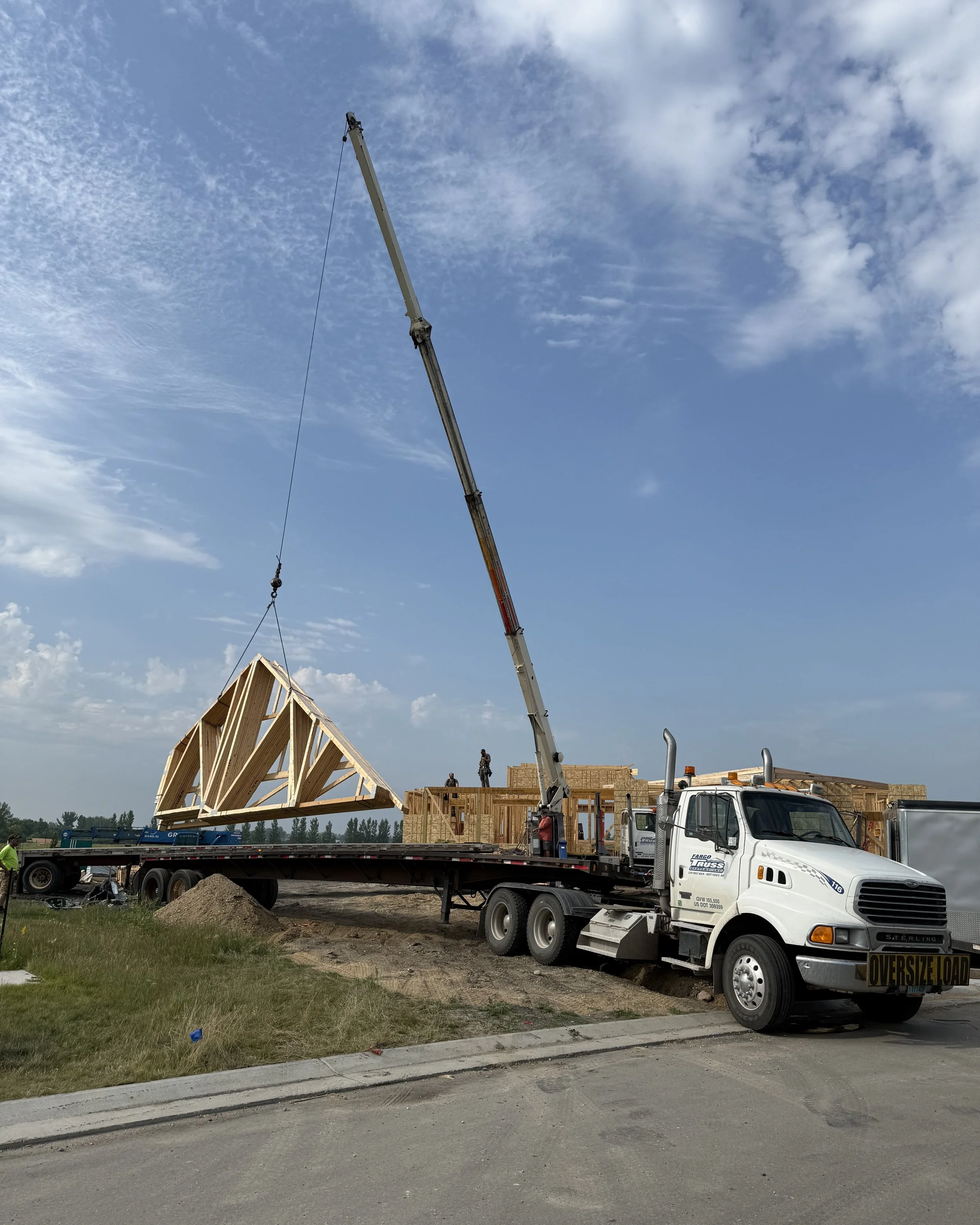 Construction site with workers and a crane lifting a wooden roof framework. A truck with an 'Oversize Load' sign is parked nearby, and partially constructed wooden building structures are visible against a partly cloudy sky.