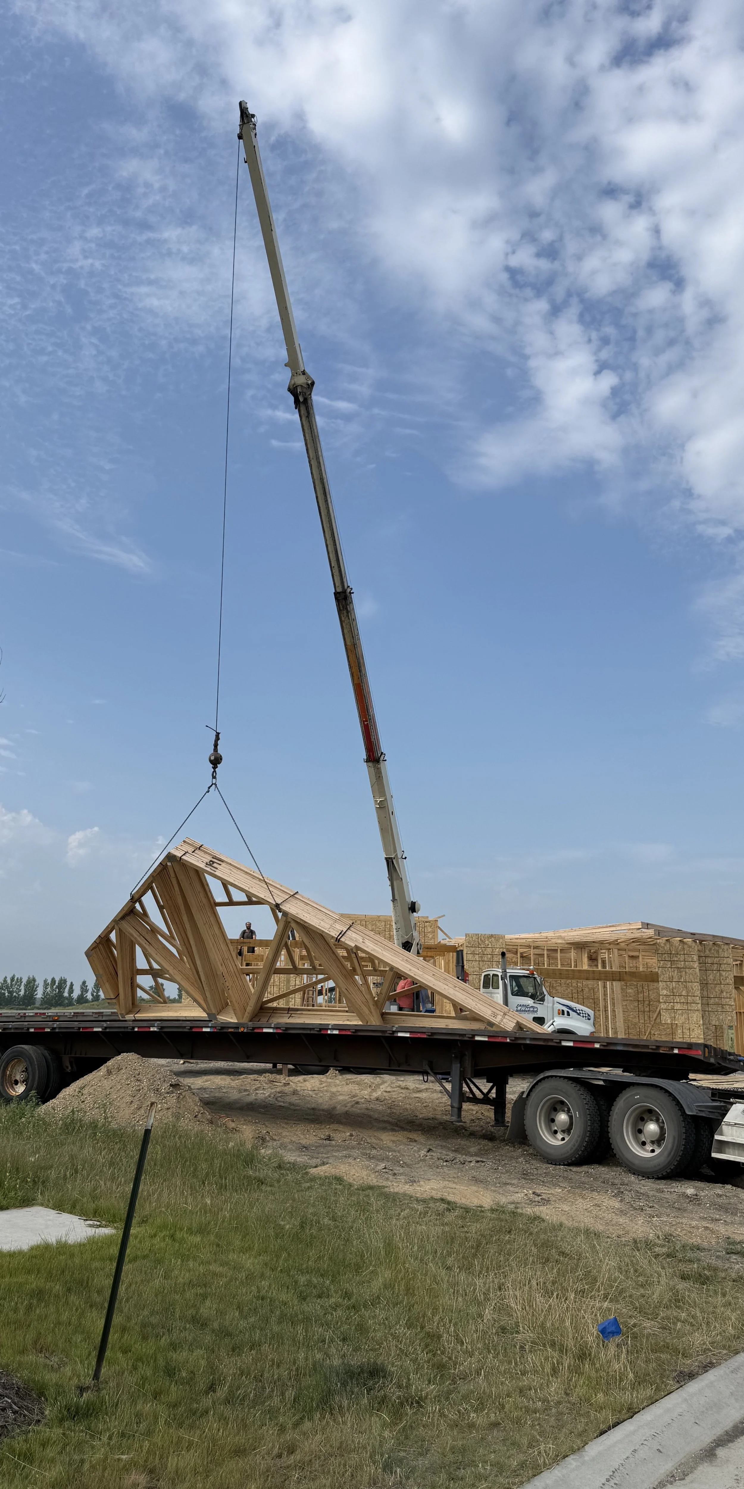 Crane lifting wooden house frame construction on flatbed truck at a construction site under a partly cloudy sky.