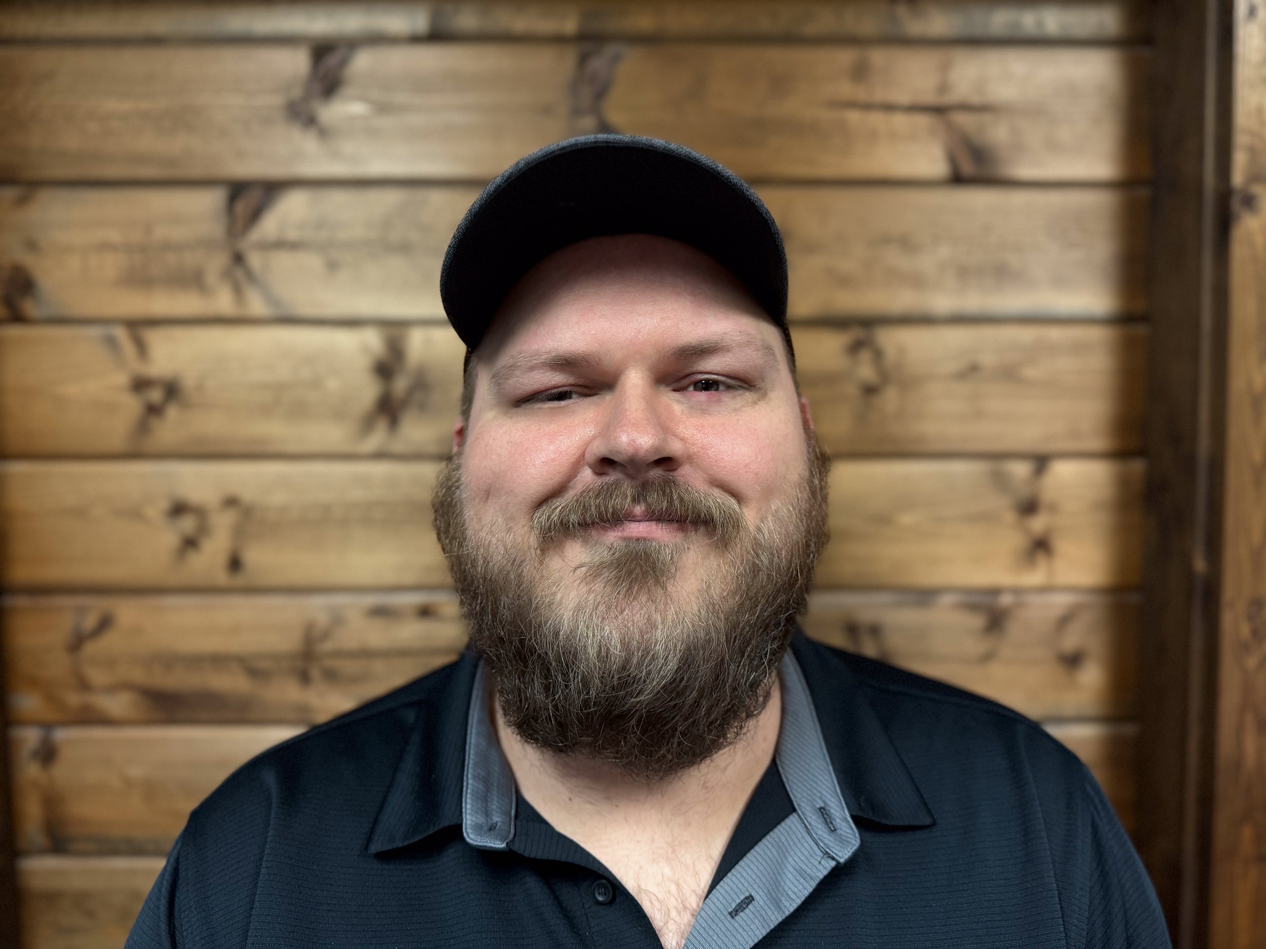A man with a beard and mustache wearing a black cap and black collared shirt, standing against a wooden wall.