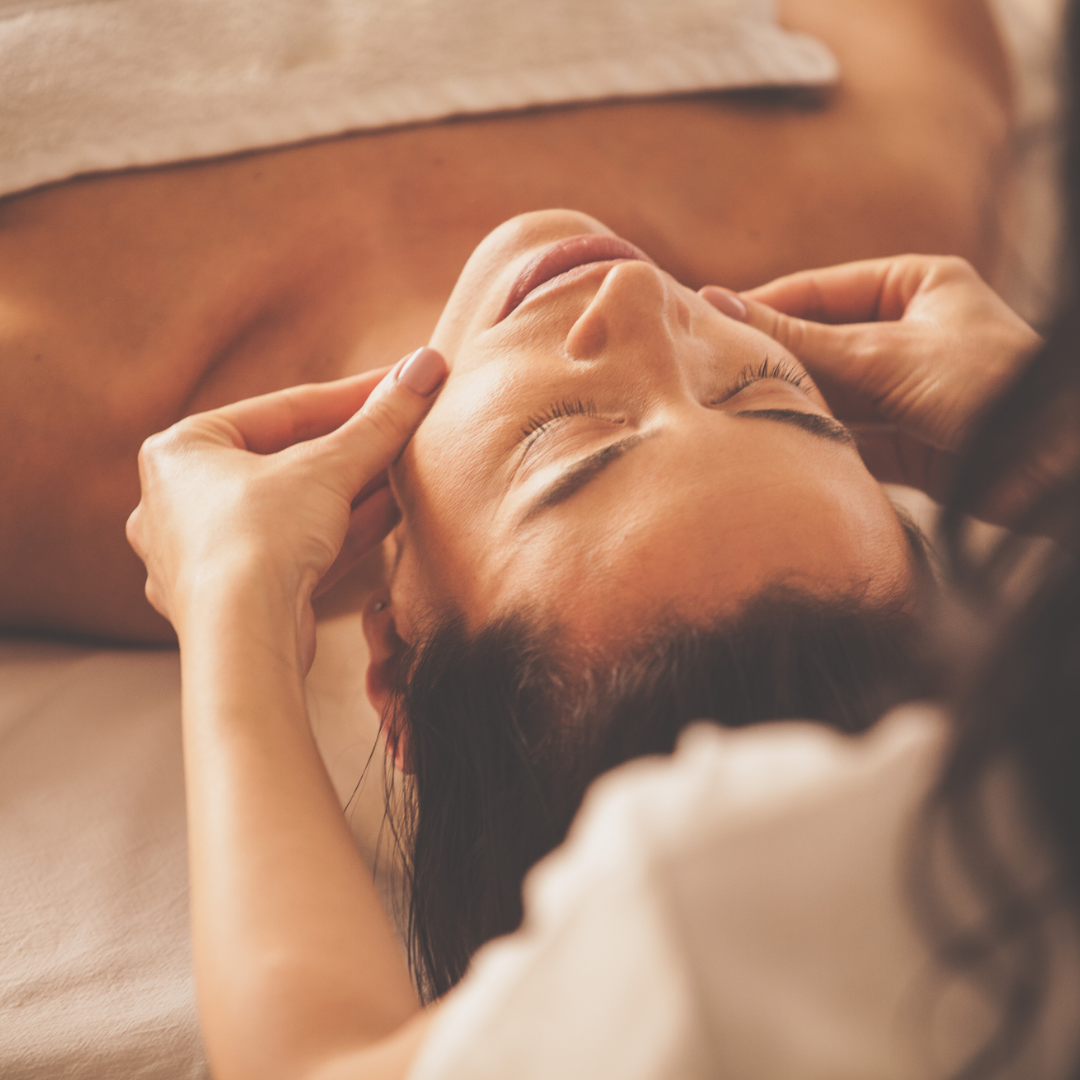 A woman receiving a facial massage with her eyes closed, lying down with a towel on her chest.