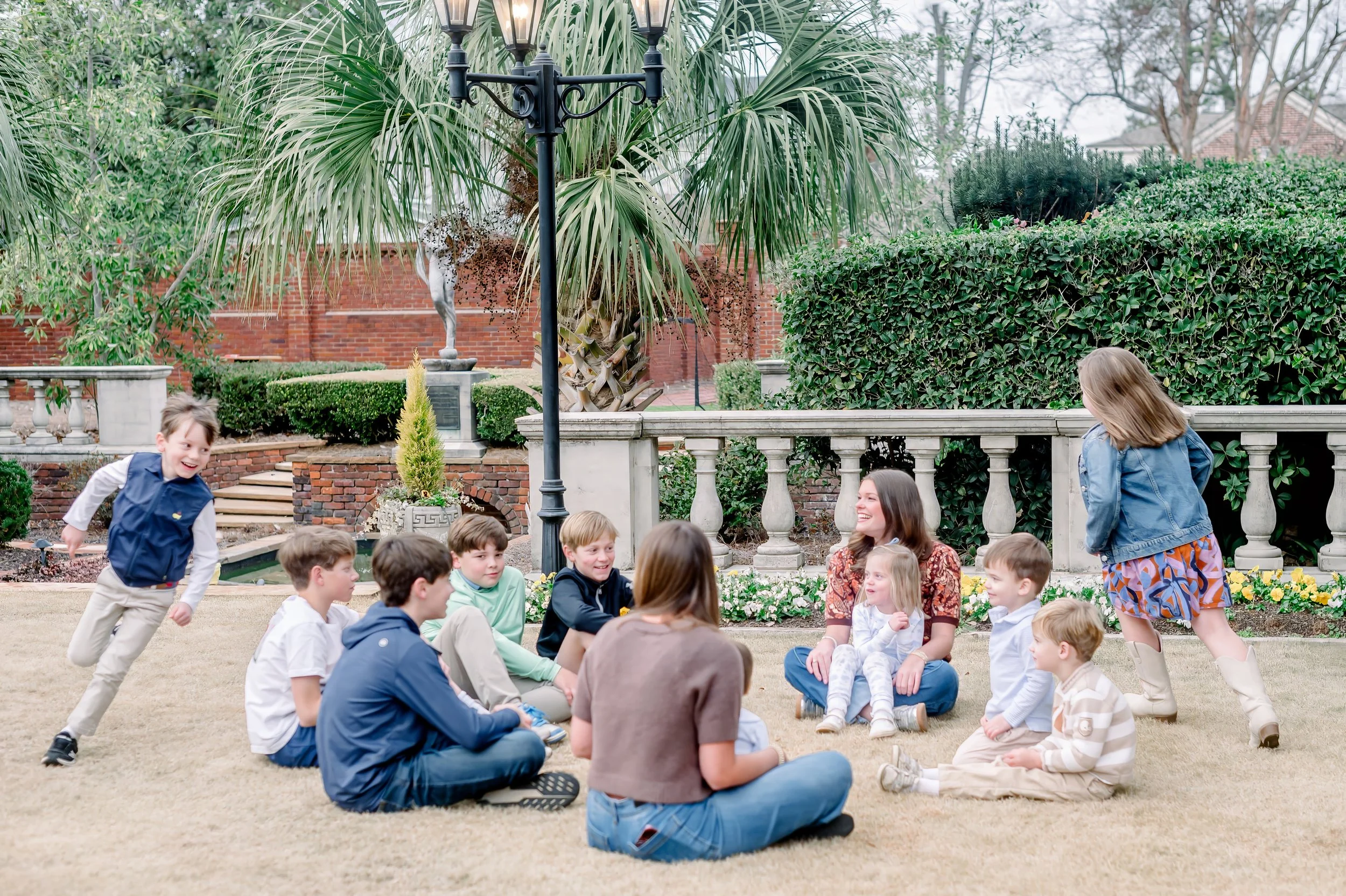 Children and two babysitters sitting and playing on the lawn in a garden with trees, bushes, flowers, a stone balustrade, and a red brick wall in the background.