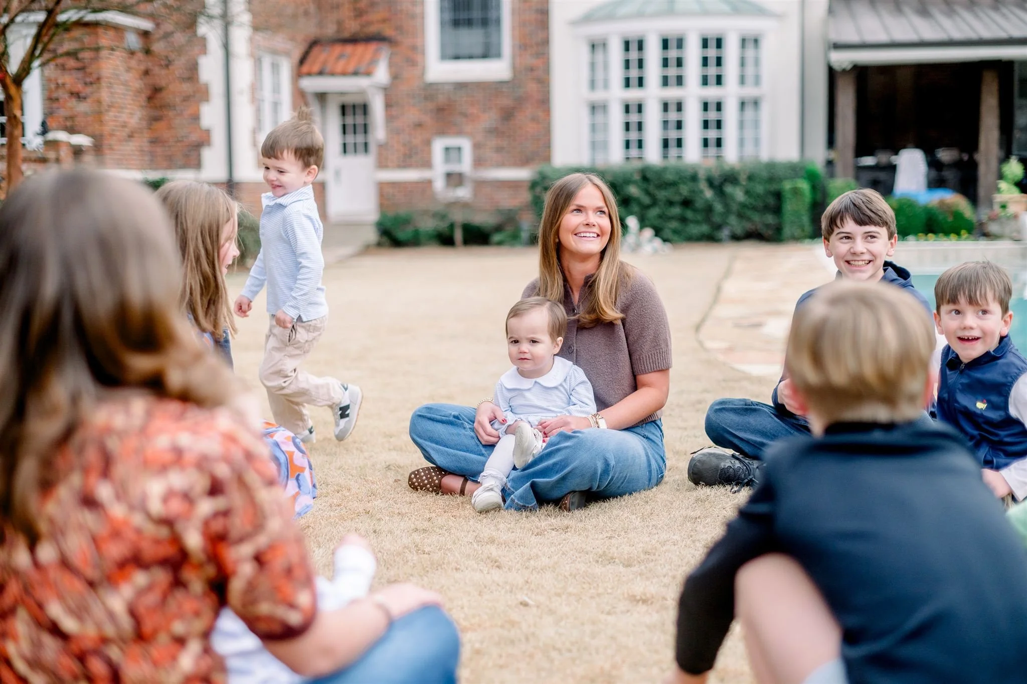 A group of children and two sitters sitting on the yard grass, smiling and enjoying outdoor activity, with a house in the background.