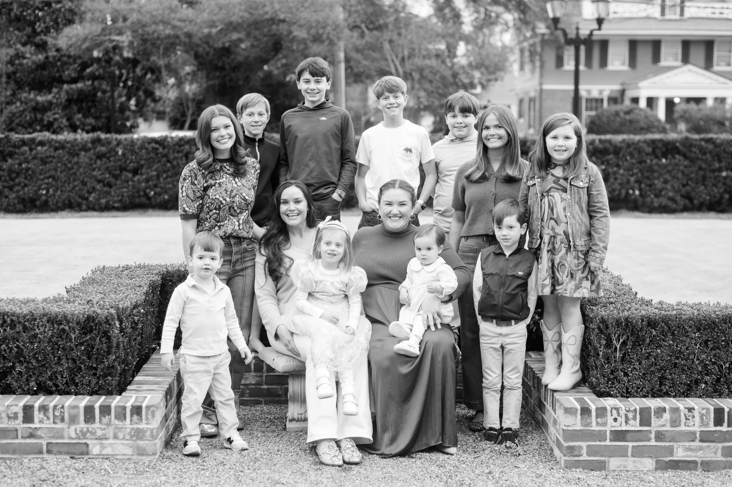 Black and white photo of a large group outdoors, with adults and children smiling, posed sitting and standing in a garden area with hedges and houses in the background.