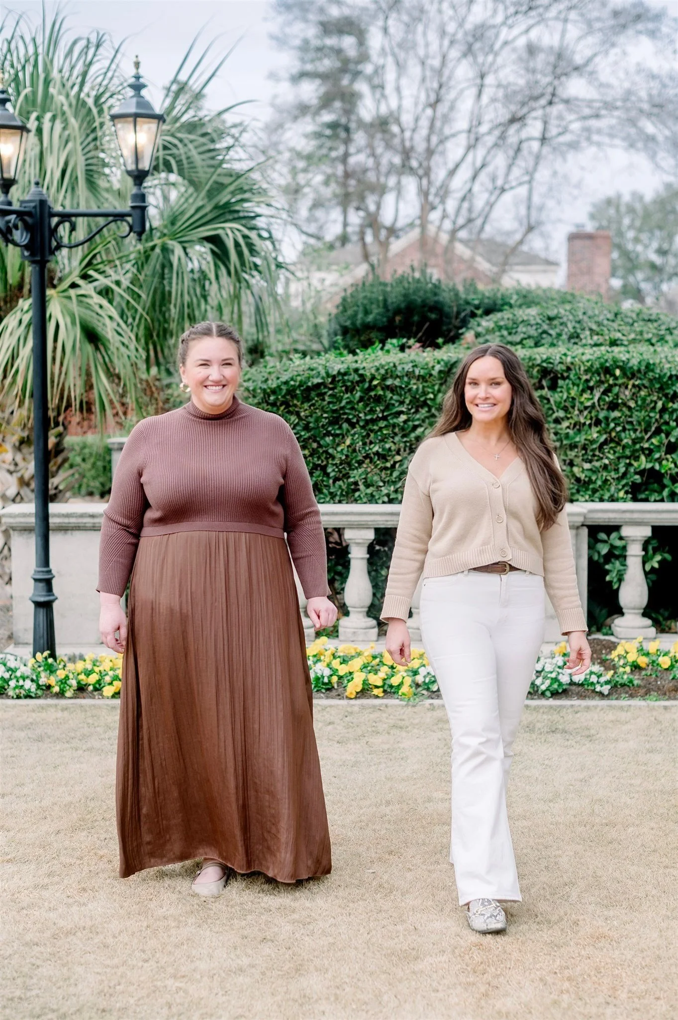 Two women walking outdoors in front of green bushes and trees, with a stone railing and colorful flowers in the background, during a cloudy day.