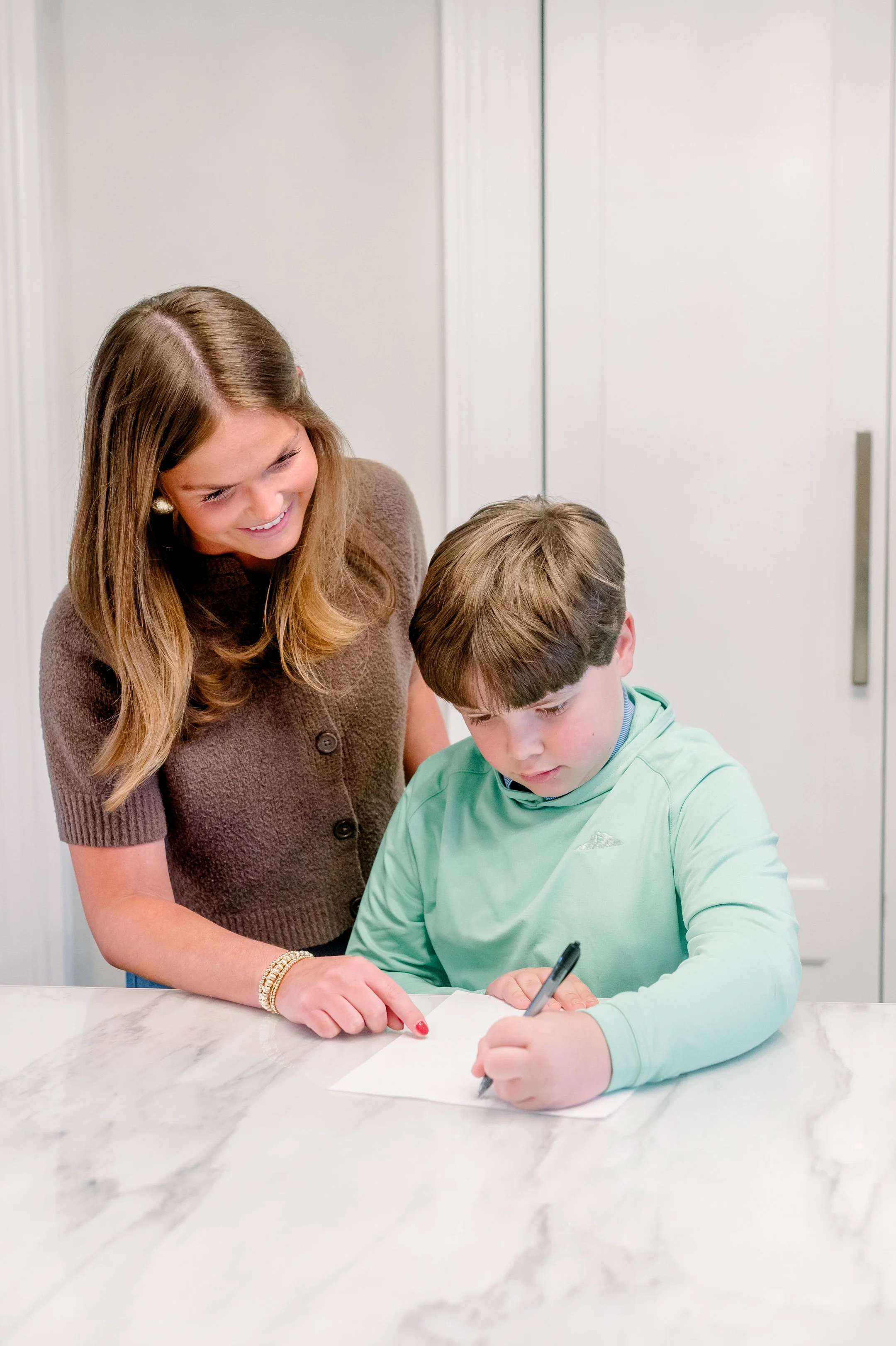 A sitter helping a young boy with homework at a white marble counter.