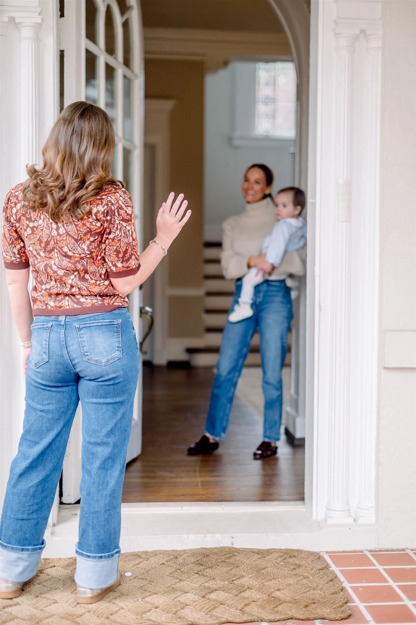 A sitter with shoulder-length hair waving at a mom holding a young child at the front door of a house.