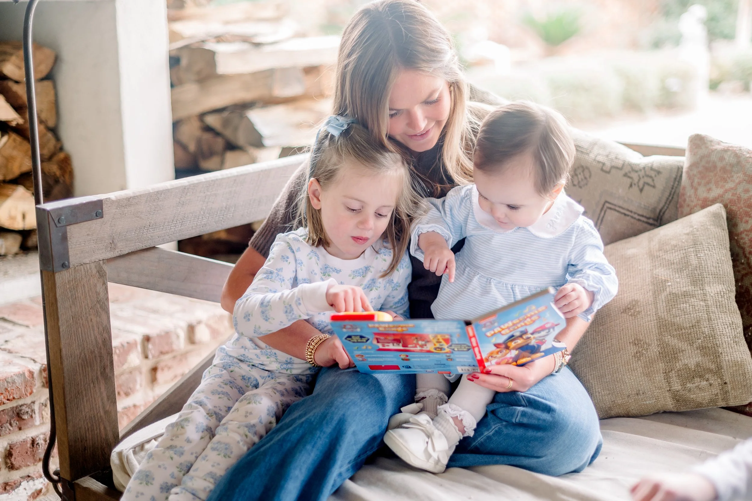 A babysitter sitting on a sofa with two young girls, all looking at a colorful children's book.