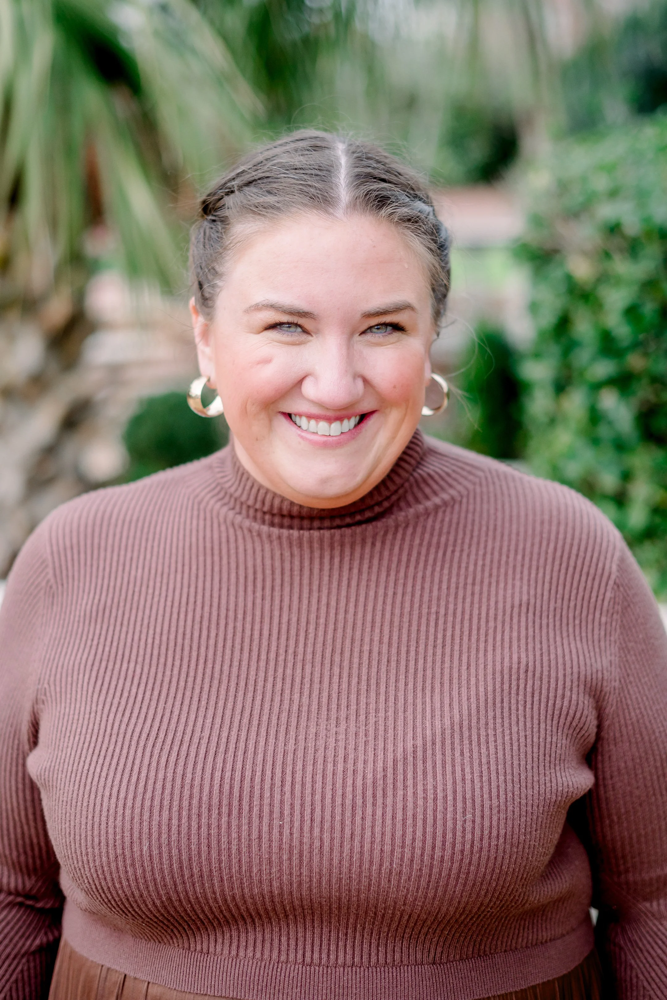 Smiling woman with braided hair and hoop earrings outdoors with green foliage in background.