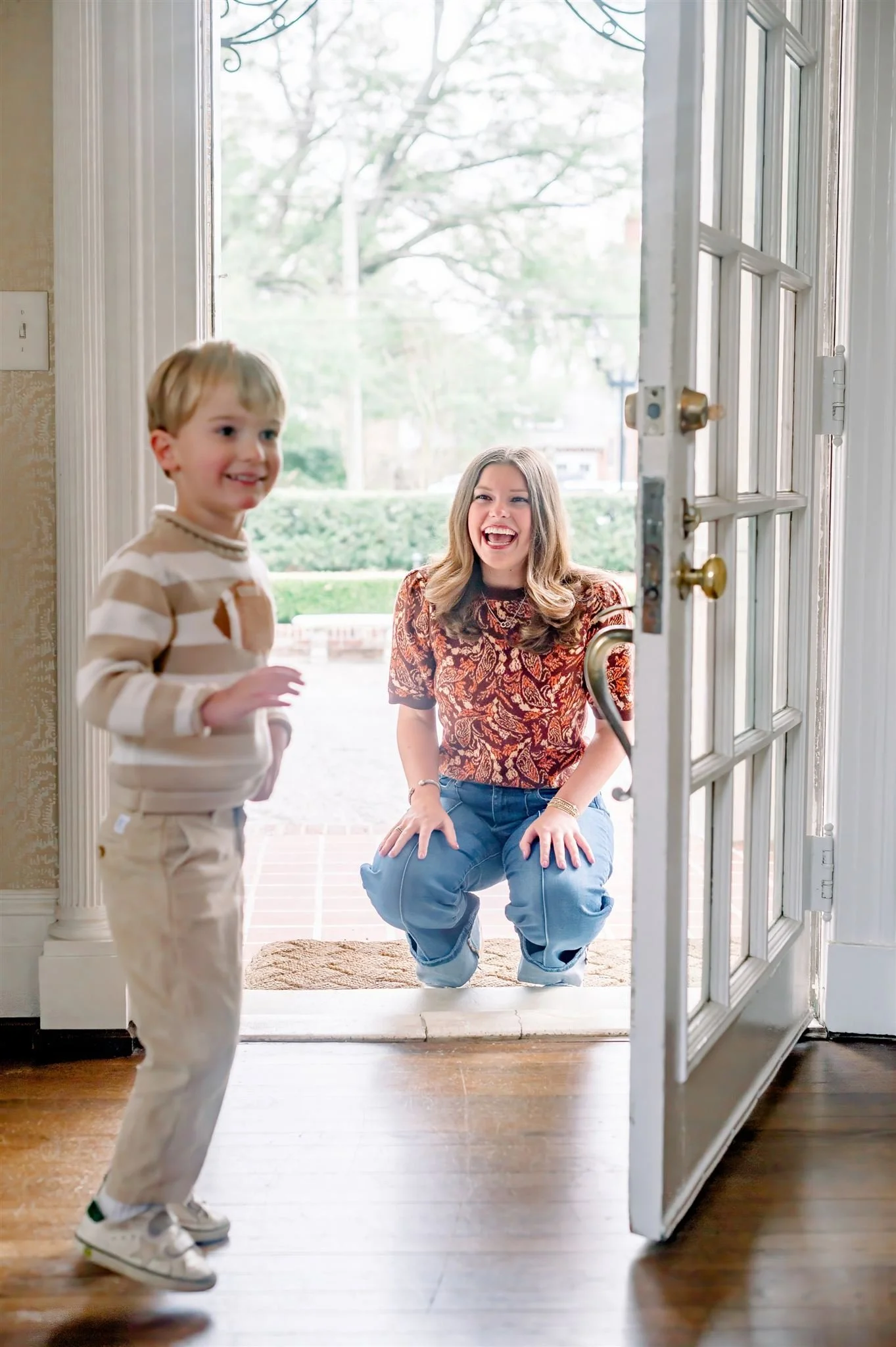 A young boy and woman are greeting each other at an open front door. The woman, with long hair, is squatting and smiling at the boy, who is walking into the house, wearing a striped sweater and beige pants.