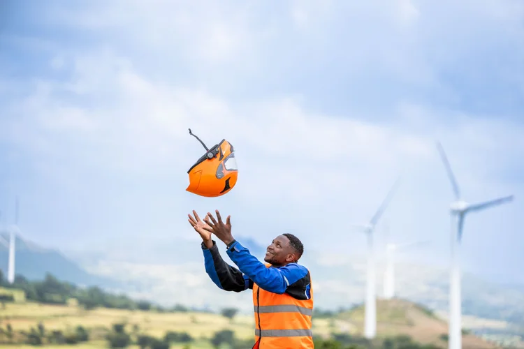 Roam rider in a blue jacket and orange safety vest throws an orange safety helmet into the air, with wind turbines in the background on a cloudy day, on the last day of the Road to Addis trip.