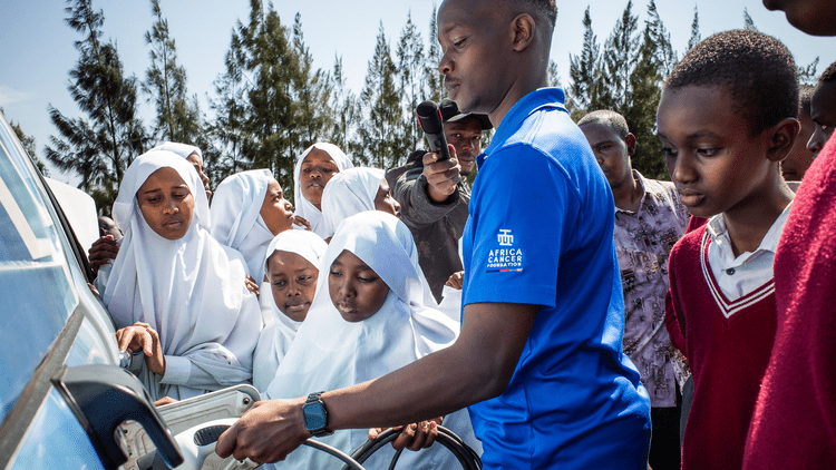 A group of children, including several girls dressed in white headscarves and white clothing, gathered around a Kenya Power technician explaining about EVs.