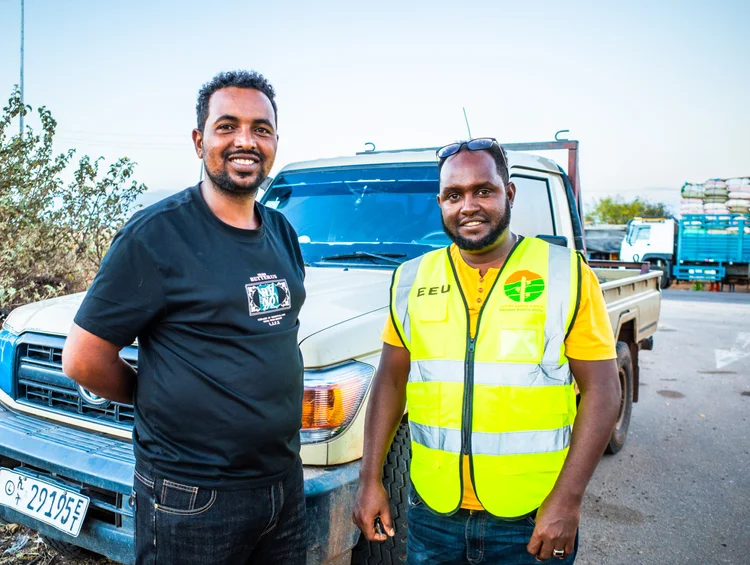 Two men from EEU (Ethiopia Electric Utility) standing in front of a pickup truck, smiling at the camera. One man is wearing a black T-shirt, and the other man is wearing a yellow safety vest with green and white details.