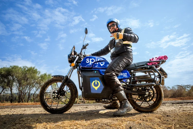 Rider on an electric Spiro motorcycle in northern Kenya on a dirt road under a blue sky, wearing a helmet, sunglasses, and motorcycle gear, making a peace sign with his hand. As part of the Road to Addis trip.