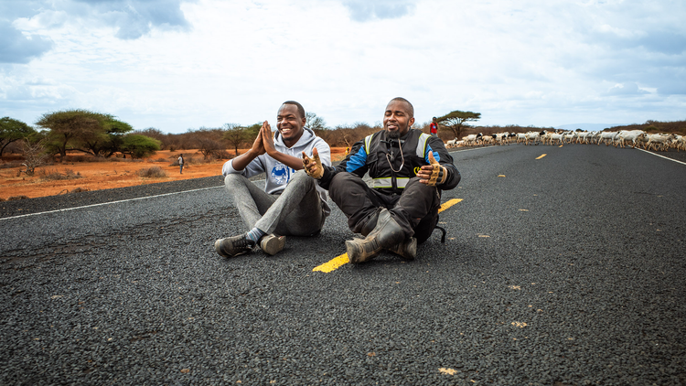 Spiro and Roam Riders on the Road to Addis, sitting on the road in northern Kenya, with a herd of cattle in the background and trees scattered across the terrain.
