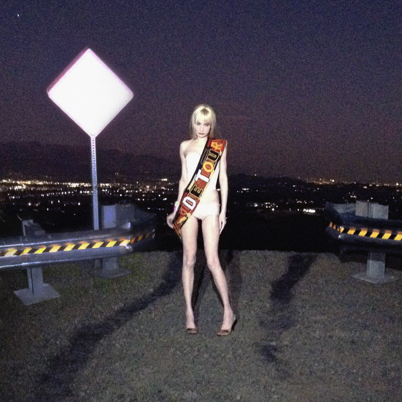 A woman with blonde hair, nude, standing on a rooftop at night near a guardrail, with city lights in the background, holding a sign that reads 'TOUR' or 'TOURIST'.