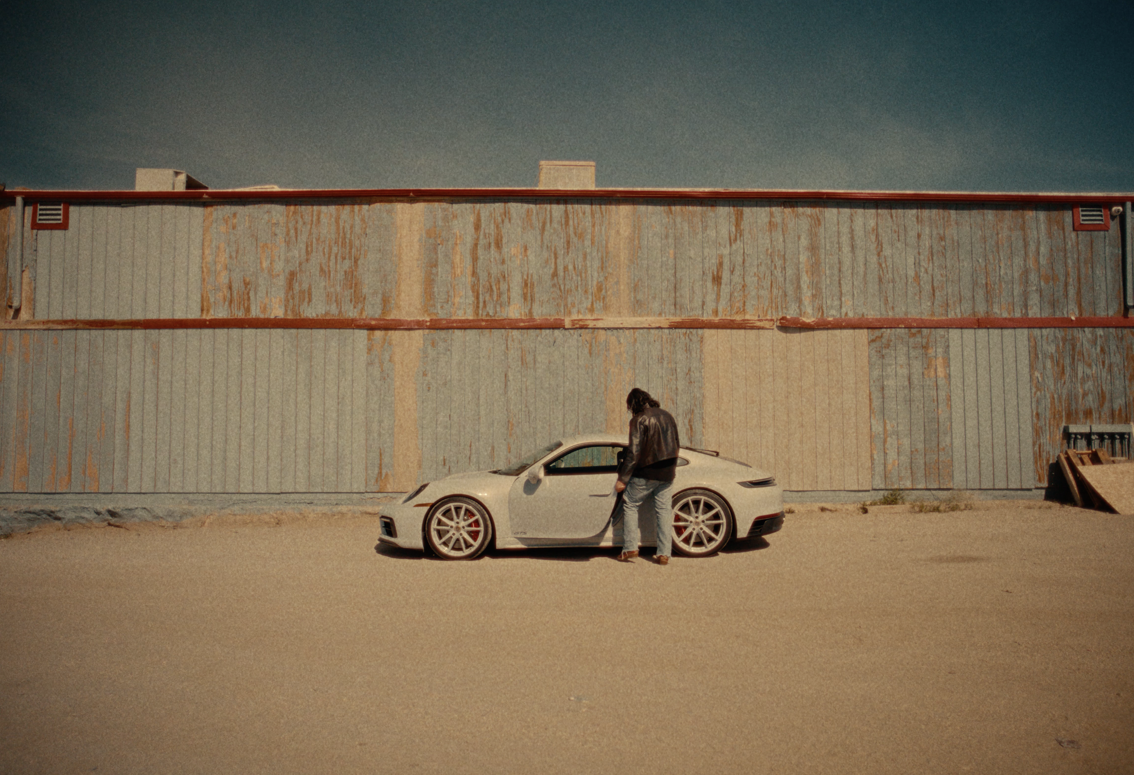 A man with long hair and a leather jacket is standing beside a white sports car in front of a weathered building with a rusted metal exterior and vents, under a clear sky.
