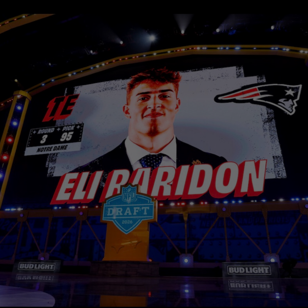 Large audience seated inside a brightly lit auditorium with multiple large screens displaying colorful graphics and text related to a sports draft event. The central screen shows the message "The Pick Is In" in neon-style lettering.
