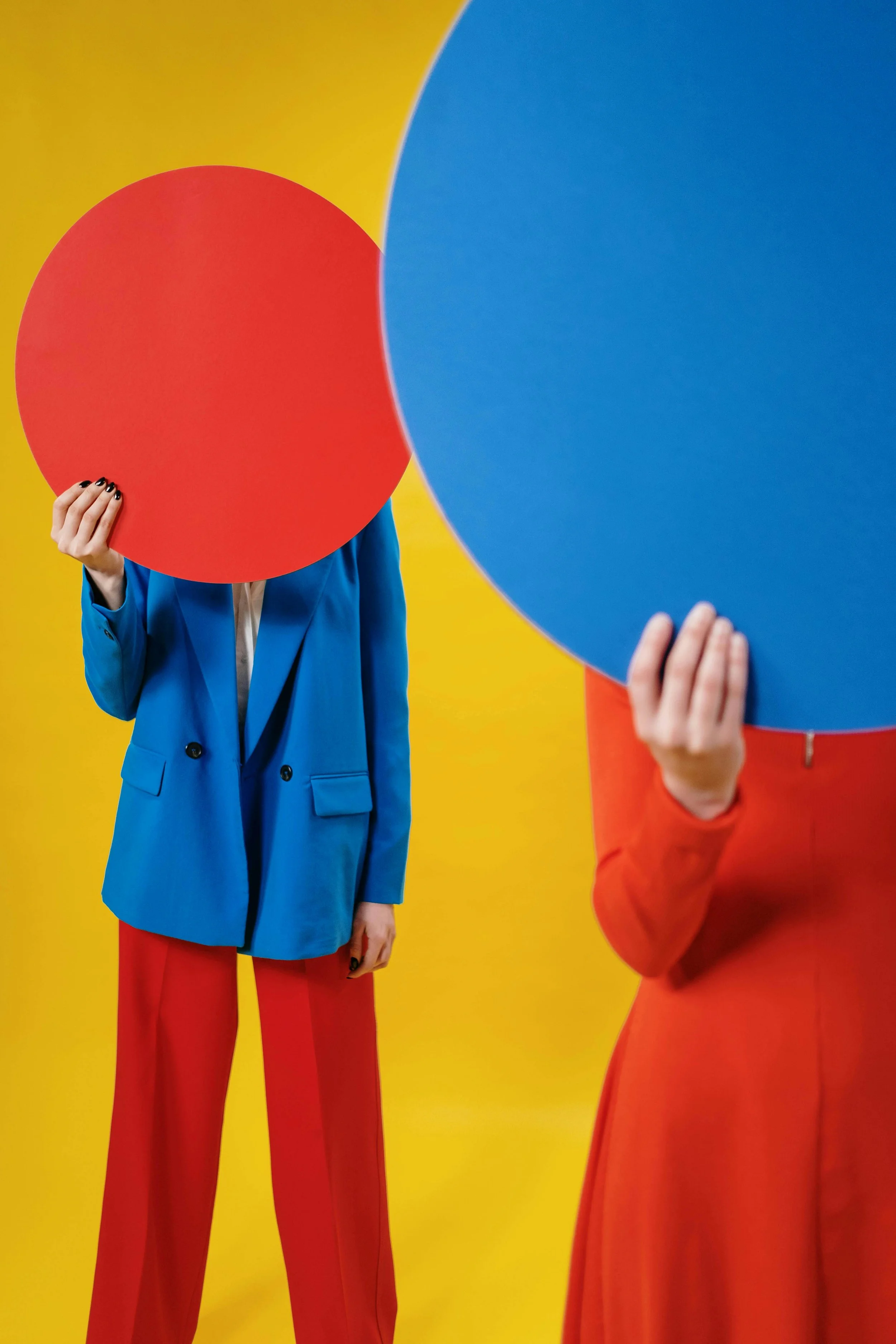 Image of two people in colorful suits holding up large circles covering their faces in opposite colors