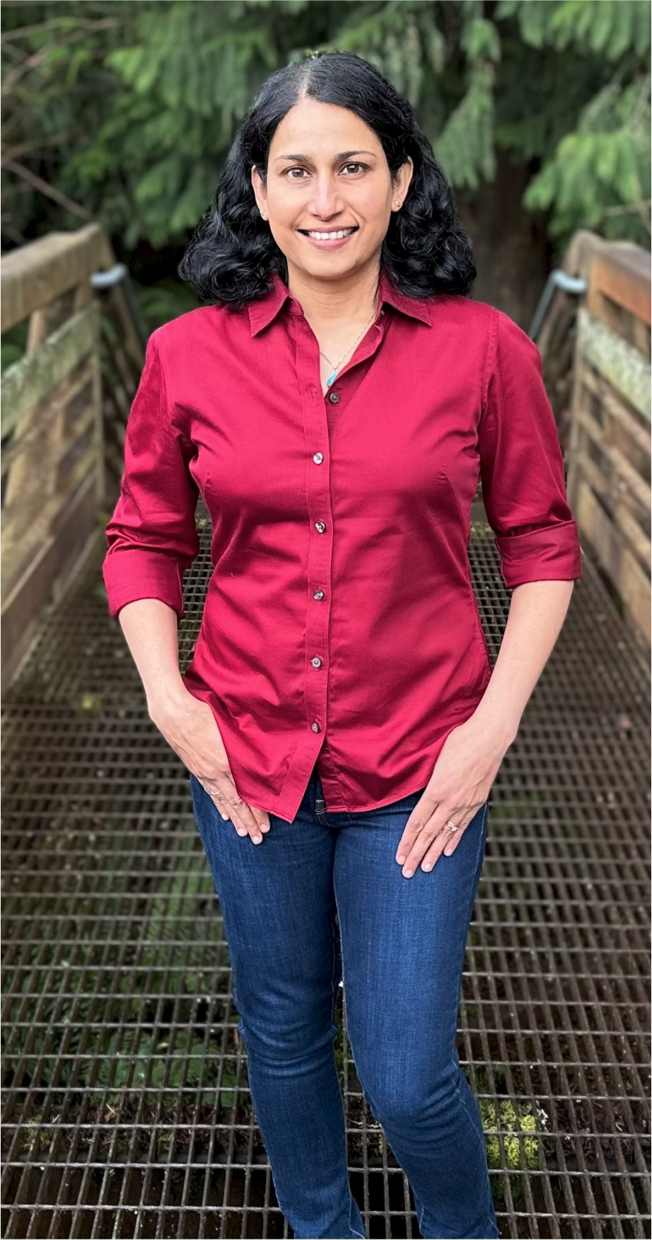 A woman with black hair wearing a red button-up shirt and blue jeans standing on a metal bridge outdoors with lush green foliage in the background.