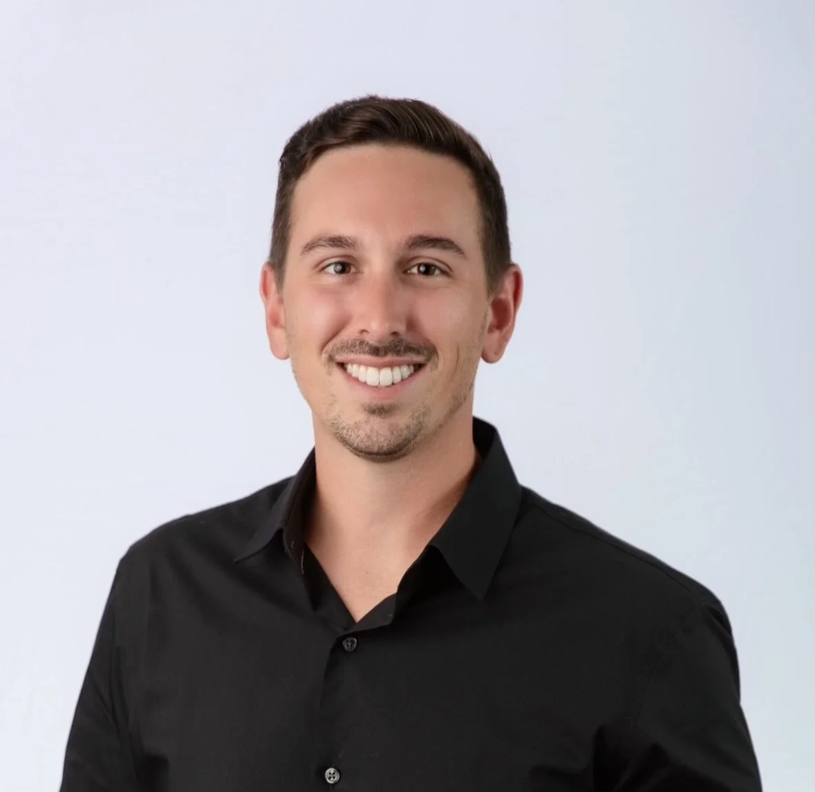 Headshot of a young man with short dark hair, smiling, wearing a black collared shirt, against a light background.