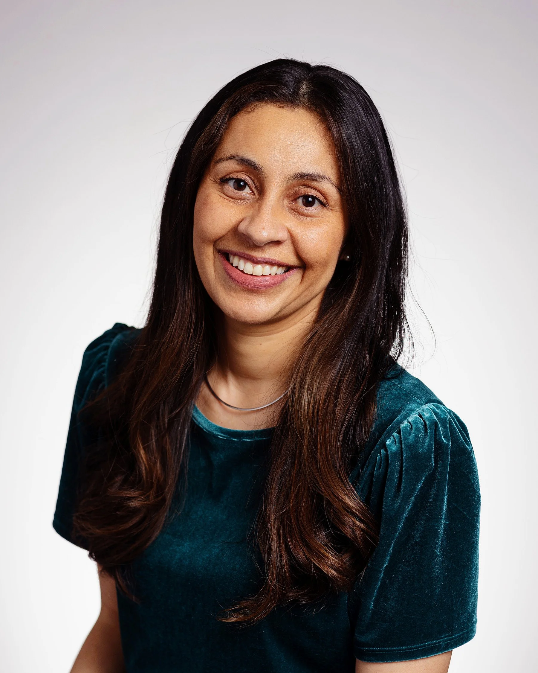 Smiling woman with long brown hair, wearing a dark teal velvet blouse, against a light gradient background.