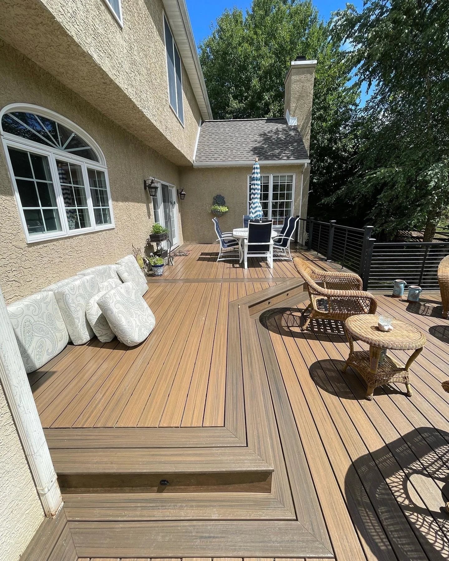 A spacious backyard deck with outdoor furniture, including a round dining table with chairs, wicker chairs, a small side table, and plants. The deck is attached to a beige house with large windows, surrounded by trees.