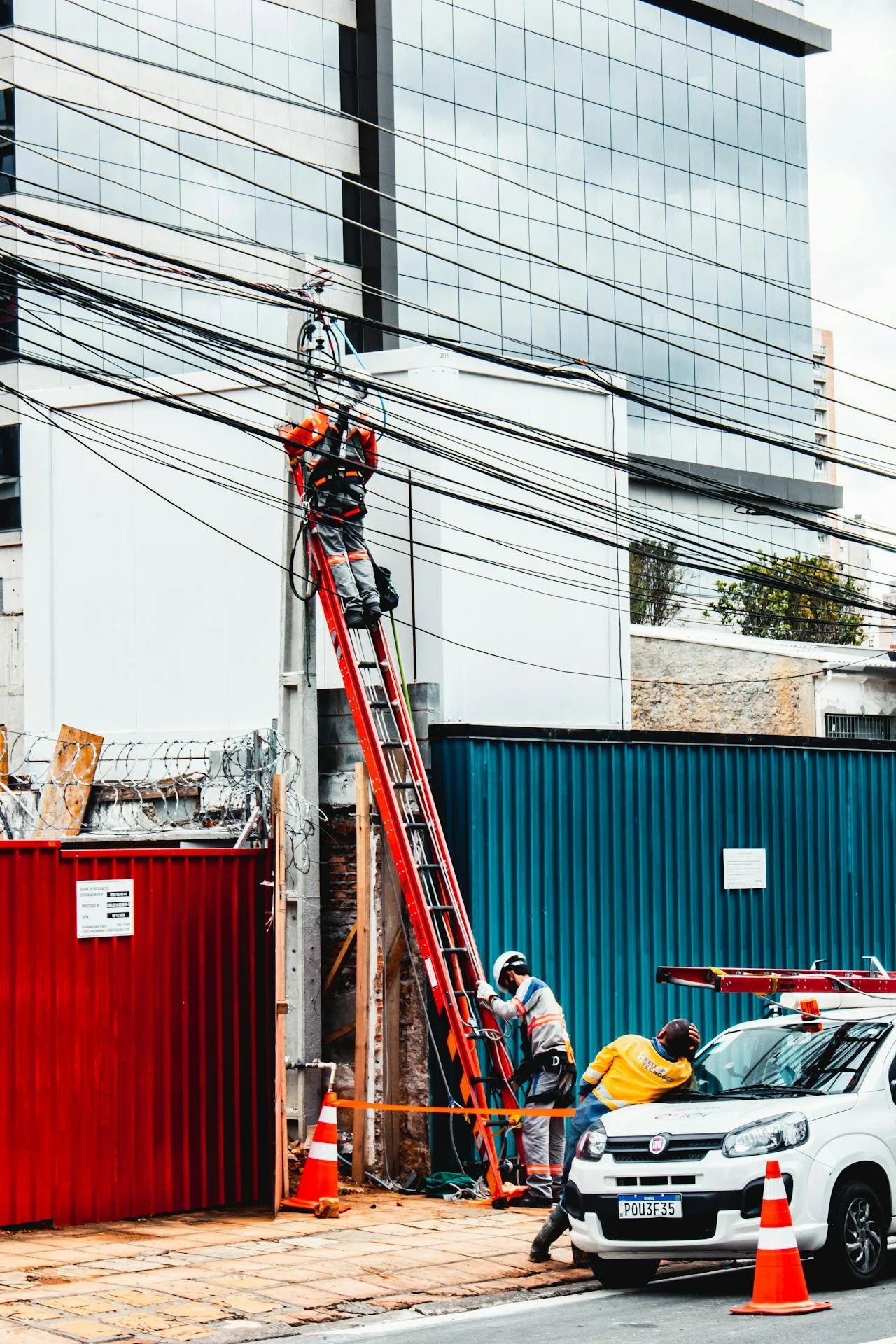 Workers repair electrical lines on a city street. One worker is on a ladder working on power lines, while others are on the ground assisting. A white car and orange traffic cones are in the foreground, with a modern glass building in the background.