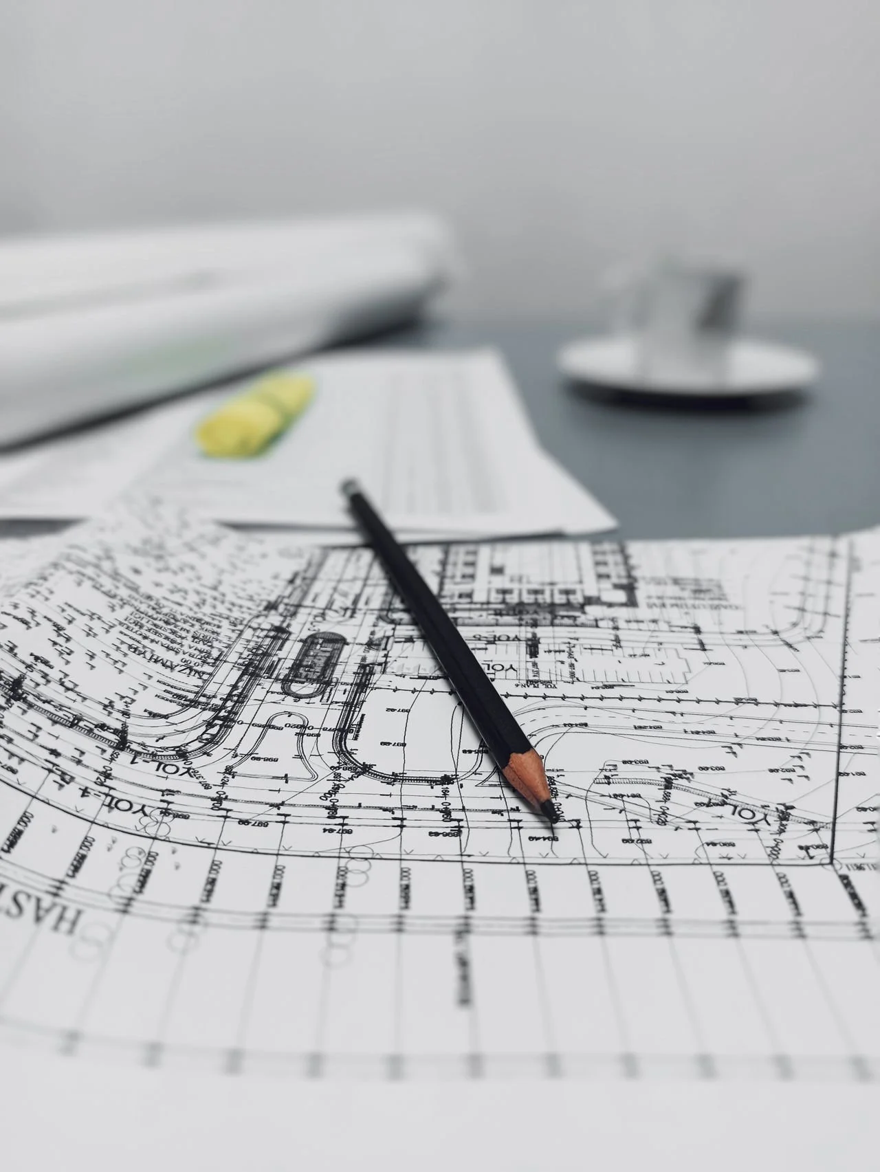 Architectural blueprint with a pencil, resting on detailed building plans, with rolled-up papers and a small dish with a framed photo in the background.