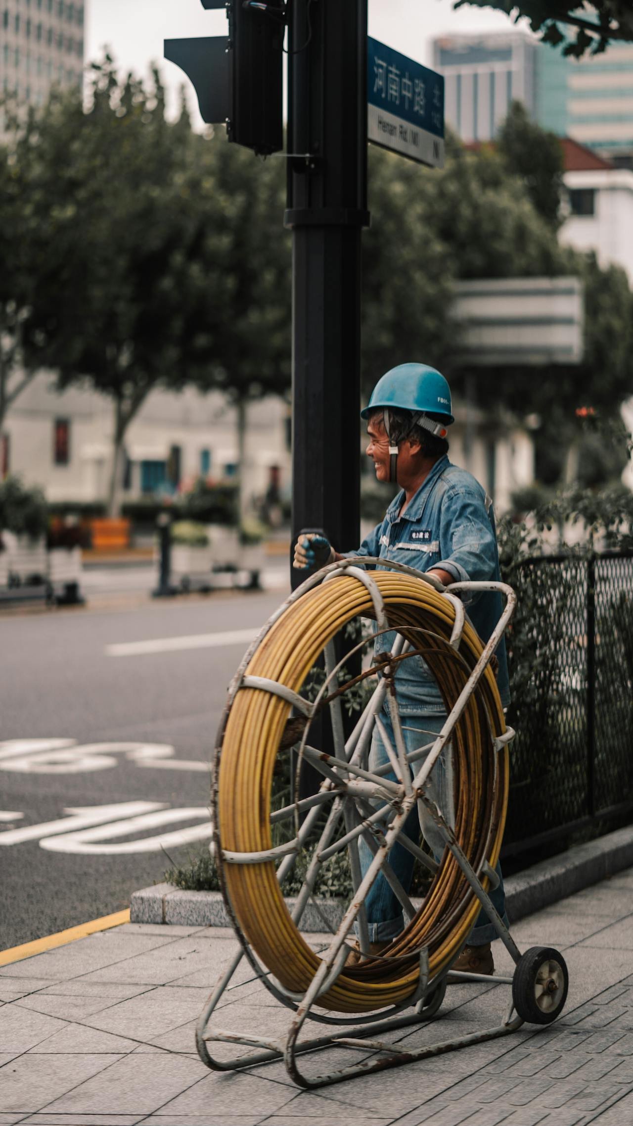 A street worker wearing a blue safety helmet and denim jacket, smiling while standing next to a cart loaded with yellow telecommunications cables, in an urban setting with trees and buildings in the background.
