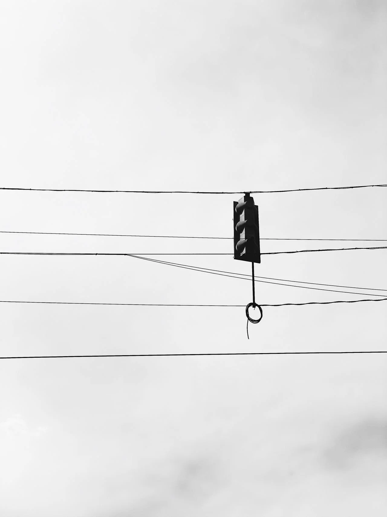 Black and white photo of a traffic light hanging from a wire against a cloudy sky, with several parallel wires crossing the frame.