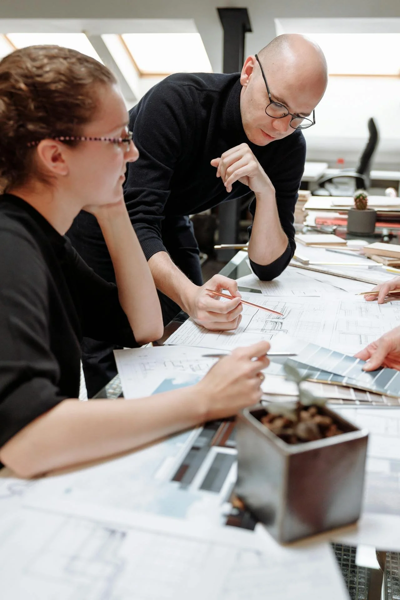 Two people working on architecture or design plans at a table with blueprints, samples, and a container of chocolates in an office setting.