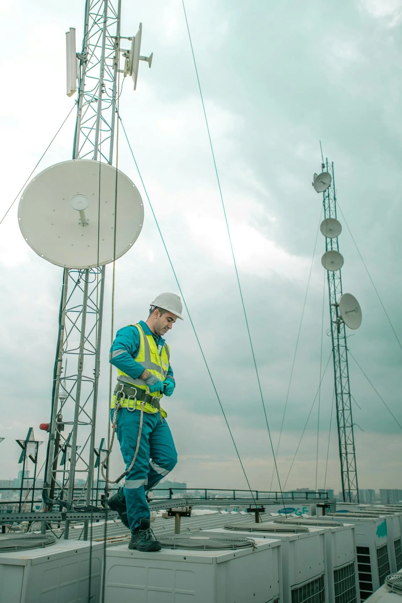 A technician wearing a white hard hat, high-visibility vest, and safety harness standing on top of an air conditioning unit at a rooftop, with communication antennas and satellite dishes in the background under an overcast sky.