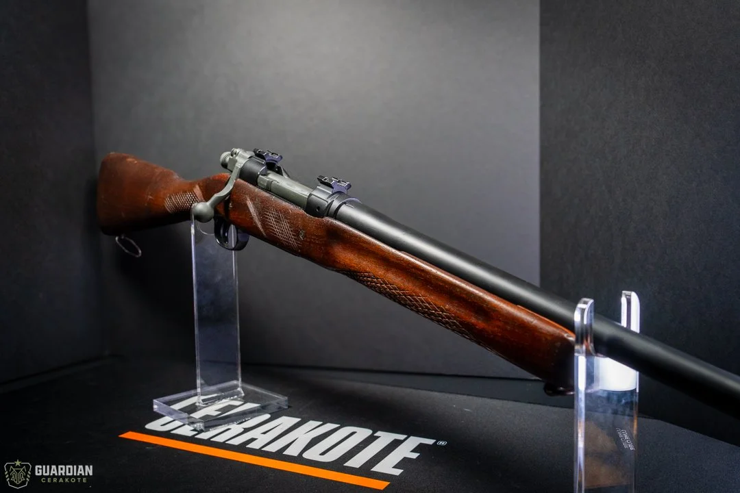 A vintage bolt-action rifle with a wooden stock displayed on a clear stand against a black background at the Guardian Cerakote exhibit.