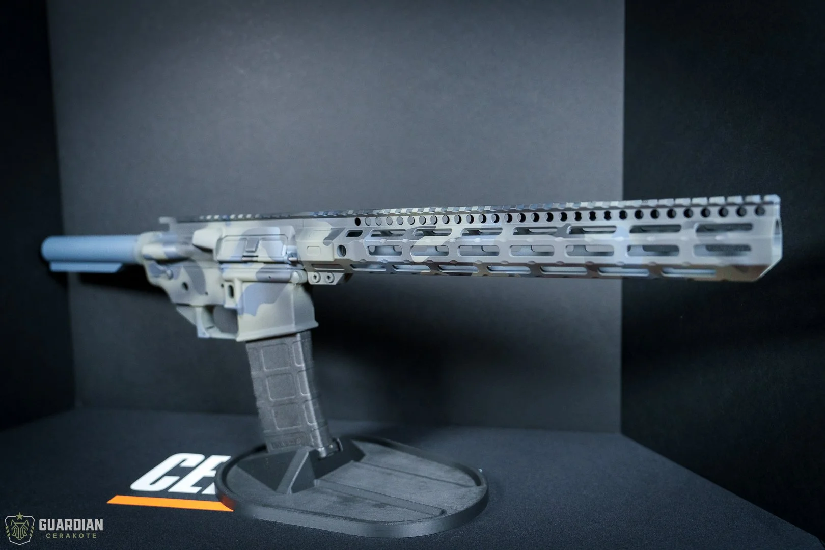 A modern rifle with a long barrel, mounted on a display stand, against a black background with logos. The rifle has a light-colored modular handguard and a transparent pistol grip.