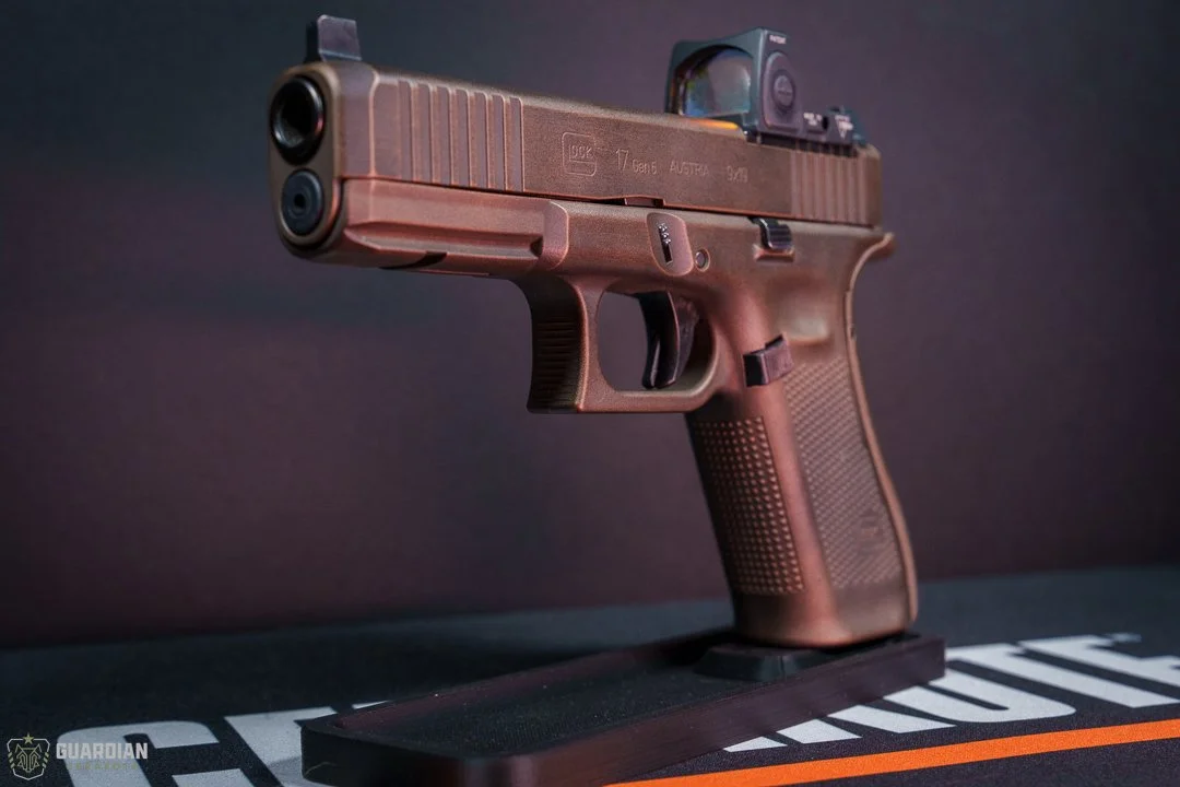 A semi-automatic pistol with a brown finish, equipped with a mounted optic sight, placed on a black display stand.