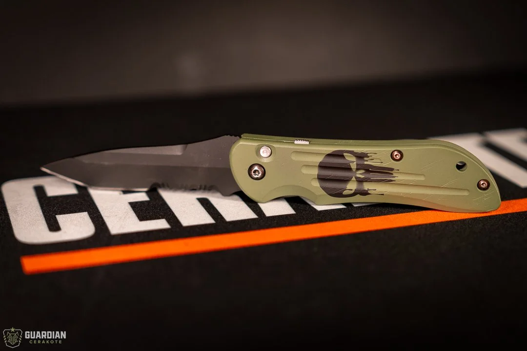 A folding knife with a black blade and an olive green handle featuring a black skull logo, resting on a black surface with the word Guardian CEKAKOTE visible.