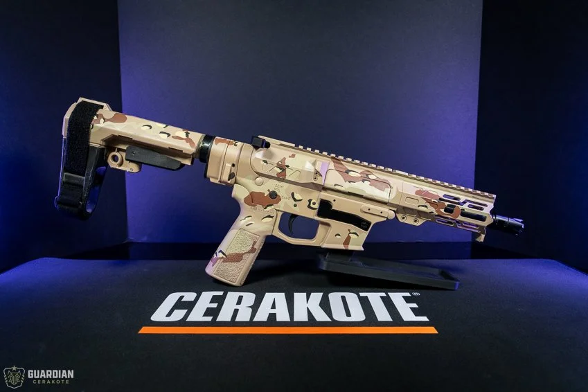 A camo-colored rifle displayed on a table with 'CERAKOTE' branding, with a dark backdrop.