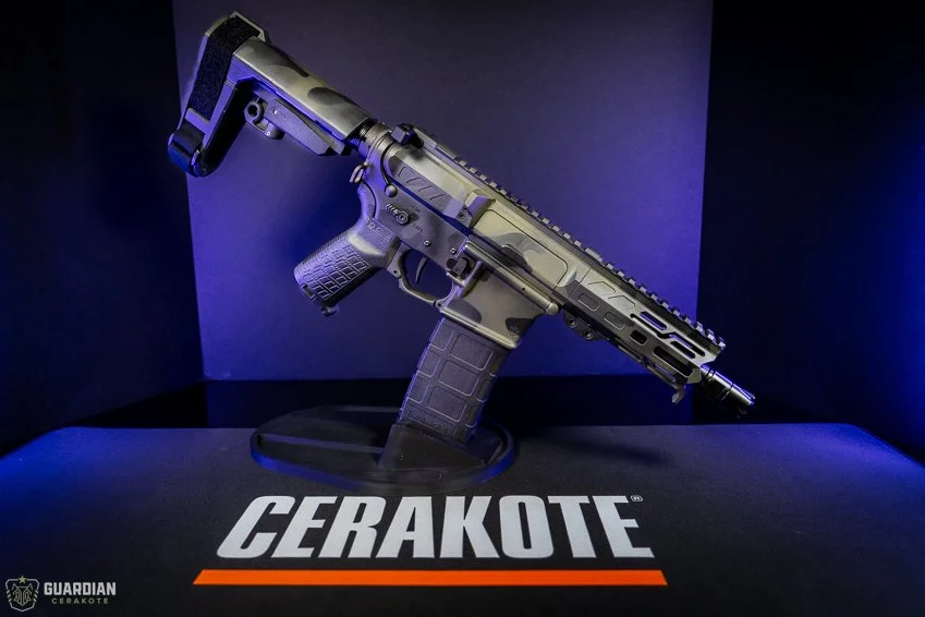 A firearm with a tan and black finish displayed on a stand with a 'CERAKOTE' logo, illuminated by blue and purple lighting.