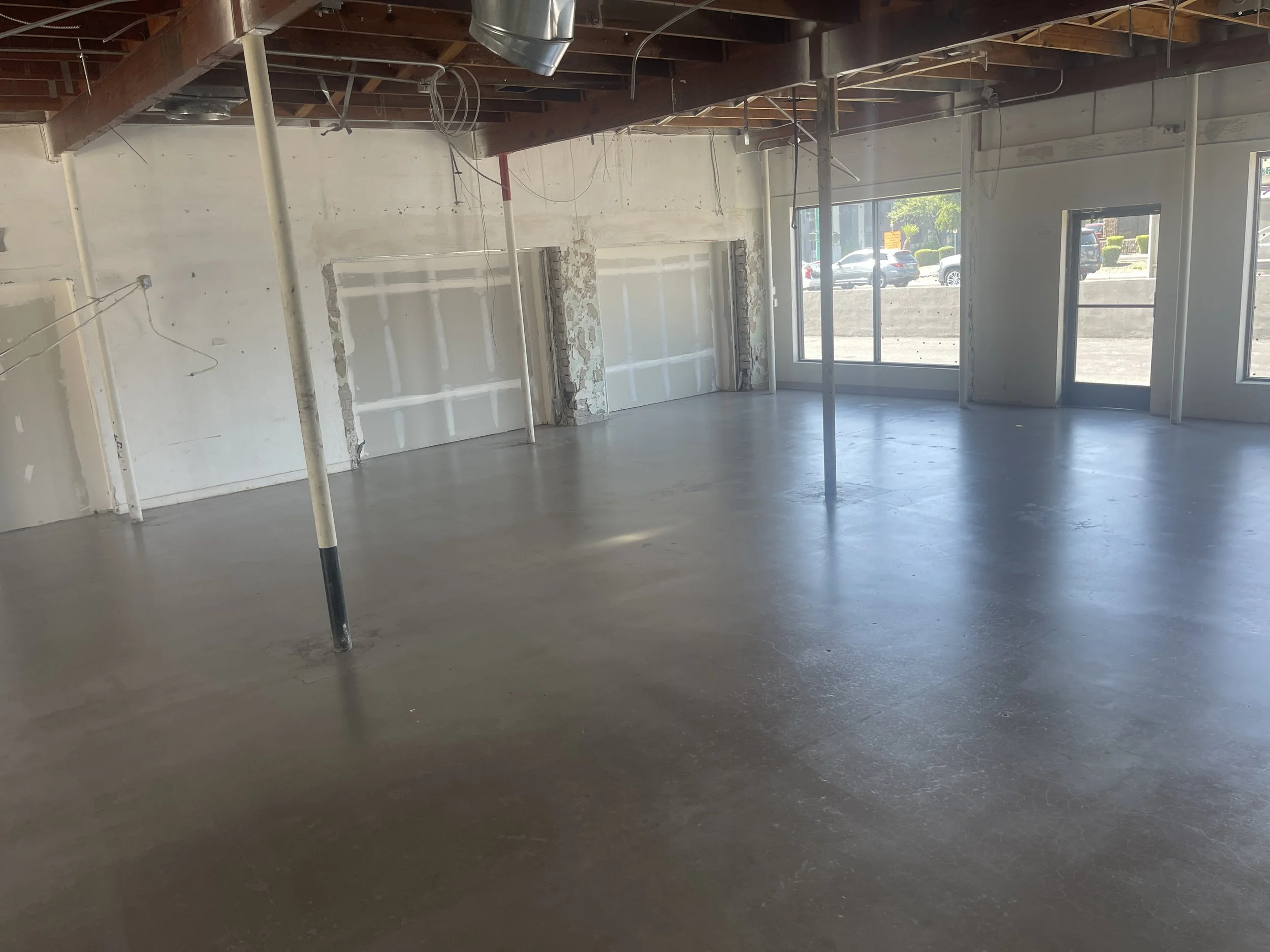 Interior of a building under construction or renovation with exposed ceiling beams, hanging wires, metal support poles, unfinished walls, and large windows letting in natural light.