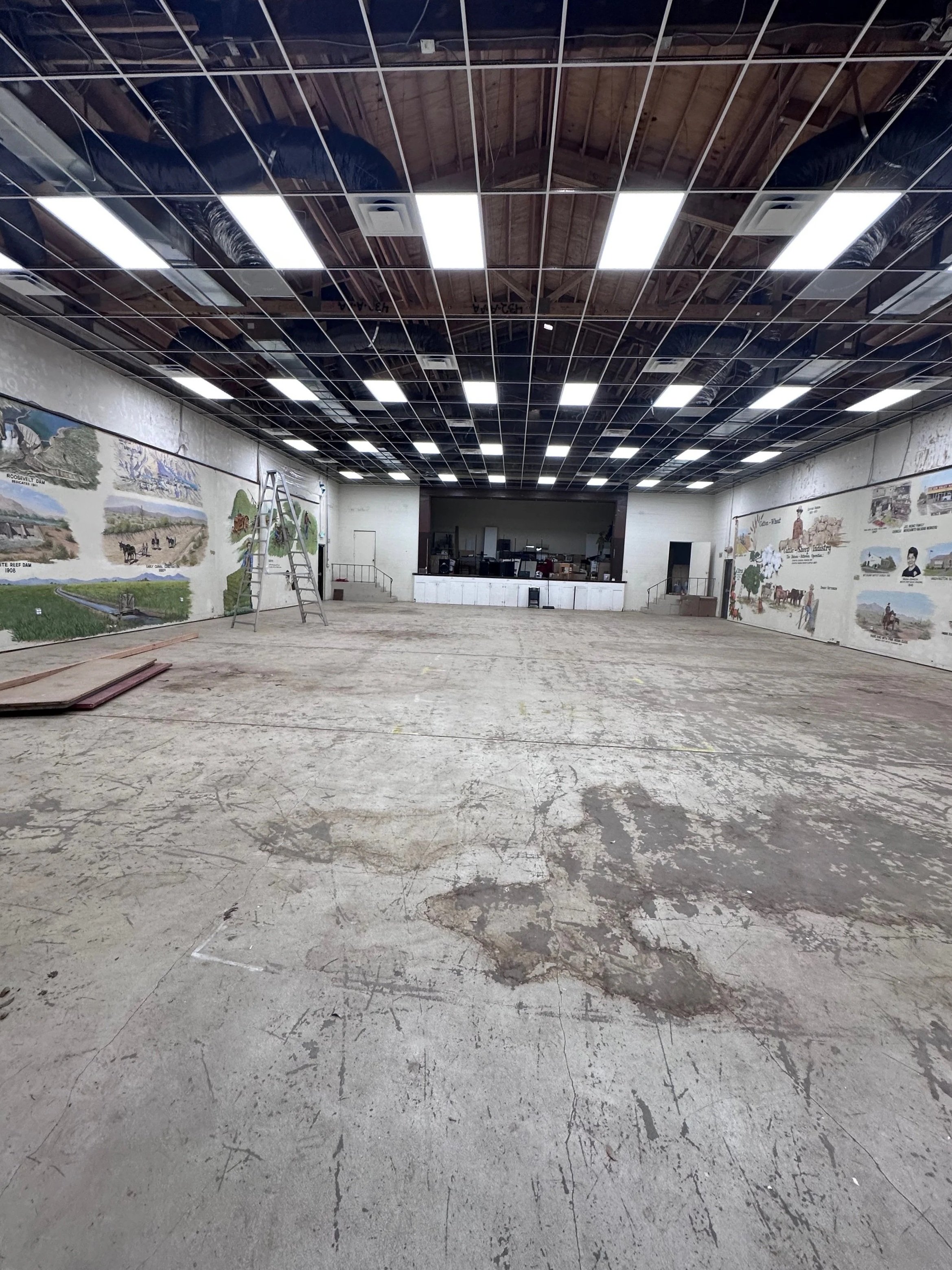 Interior view of a large, unfinished room with a concrete floor, mural paintings on the walls, and a ceiling with exposed wooden beams and black ventilation ducts. There is a ladder standing upright and some construction materials scattered on the floor.