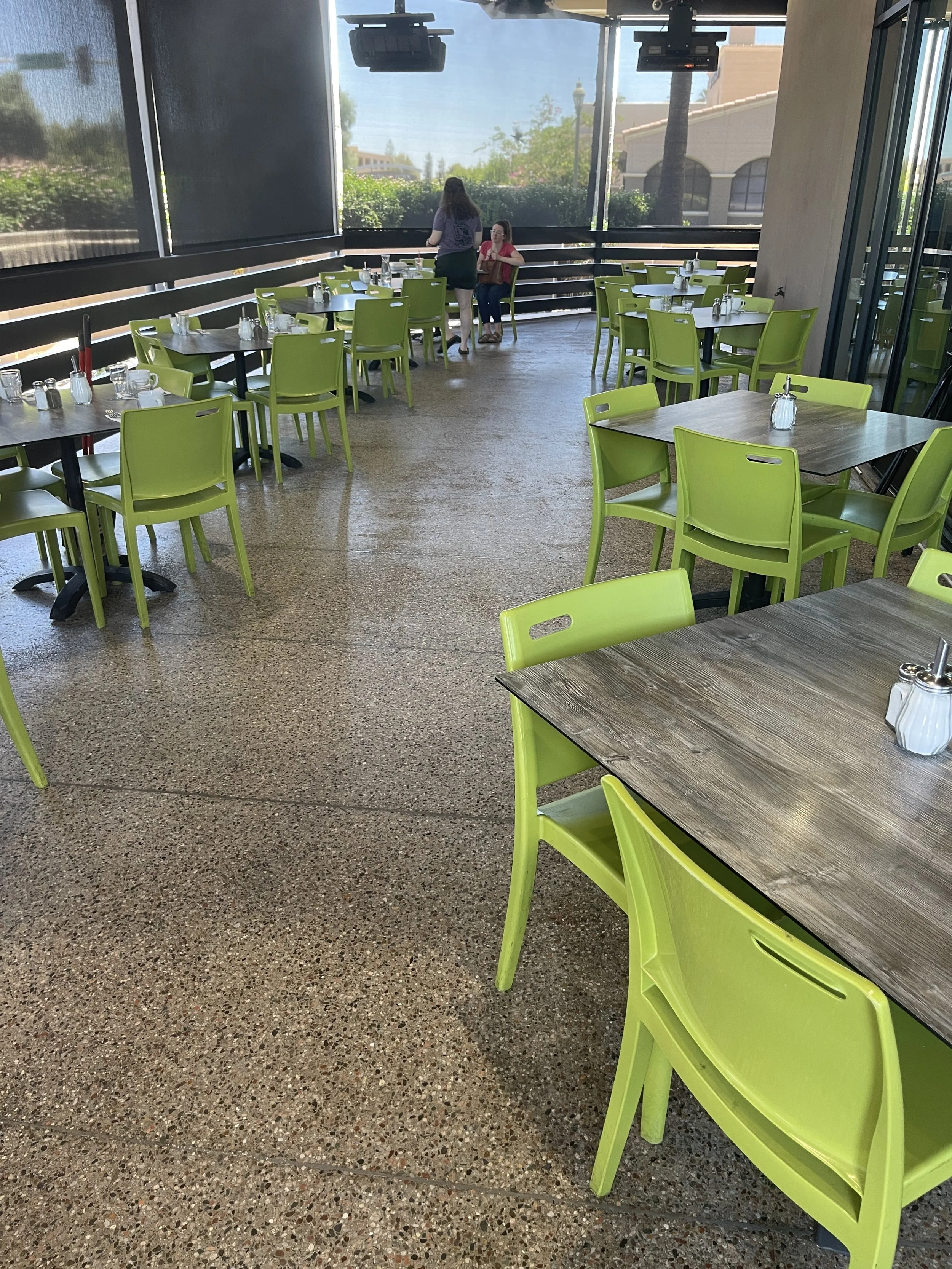 Empty restaurant with bright green chairs and wooden tables, a few patrons near the back, large windows with a view of trees and buildings outside.