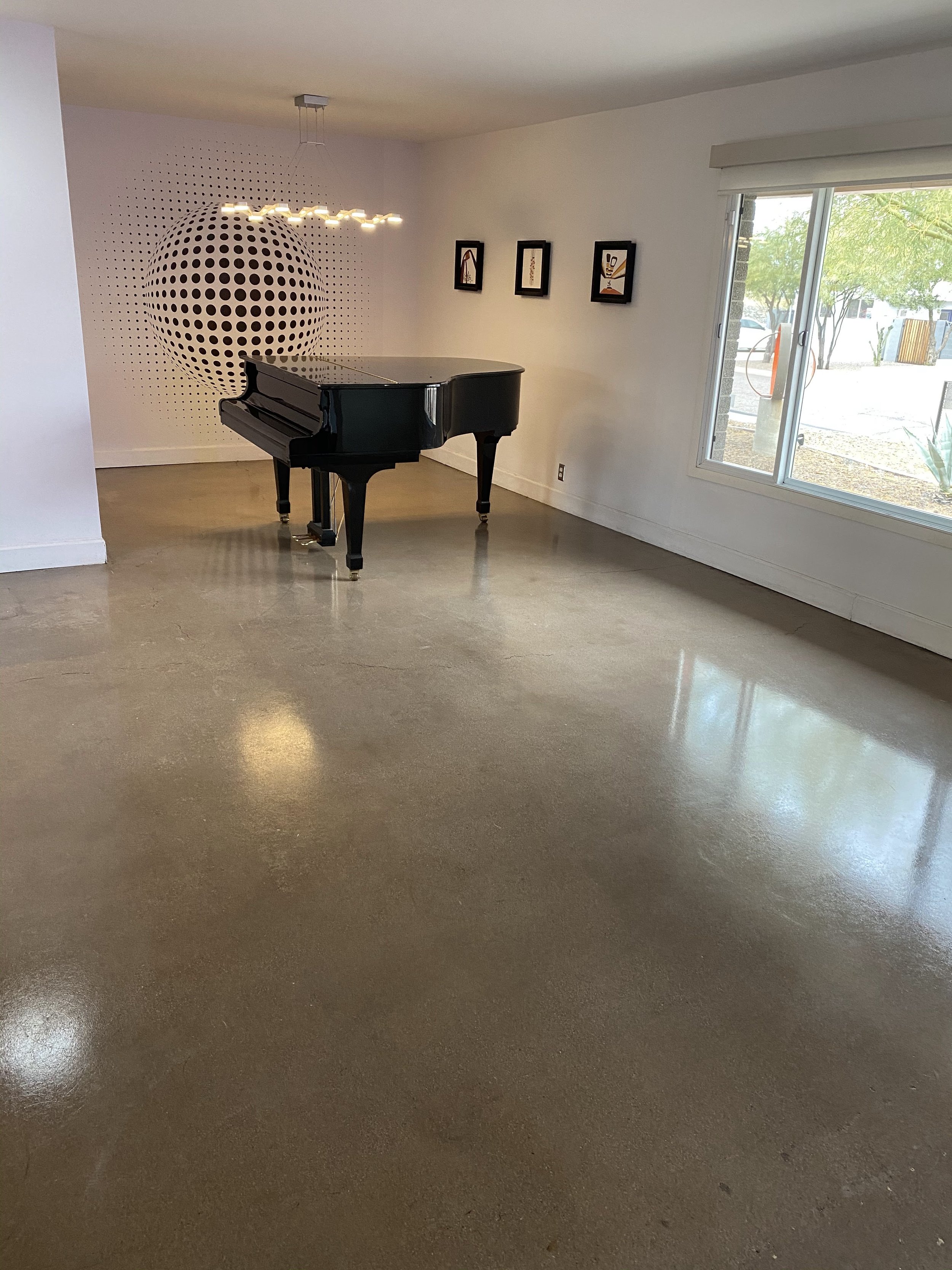 Empty room with a black grand piano, modern hanging light fixture, framed artwork on white walls, and a window showing outdoor trees and parked cars.