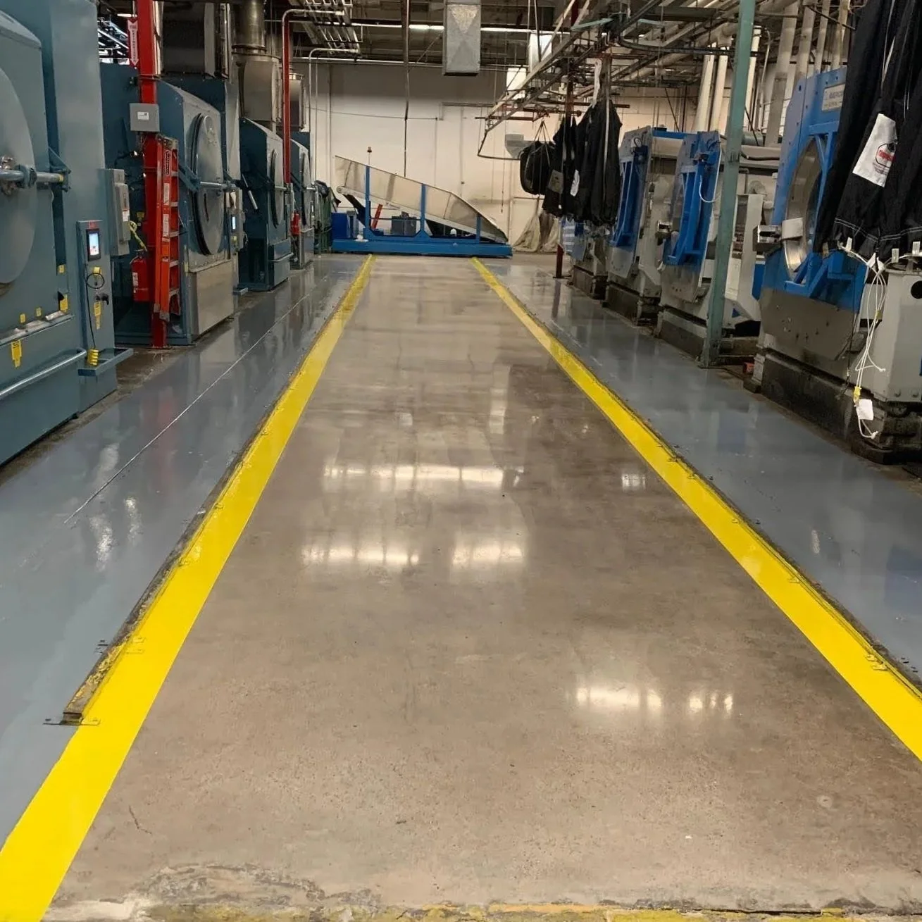 Industrial laundry room with washing machines on either side, a yellow safety line on the floor, and laundry bags hanging above.