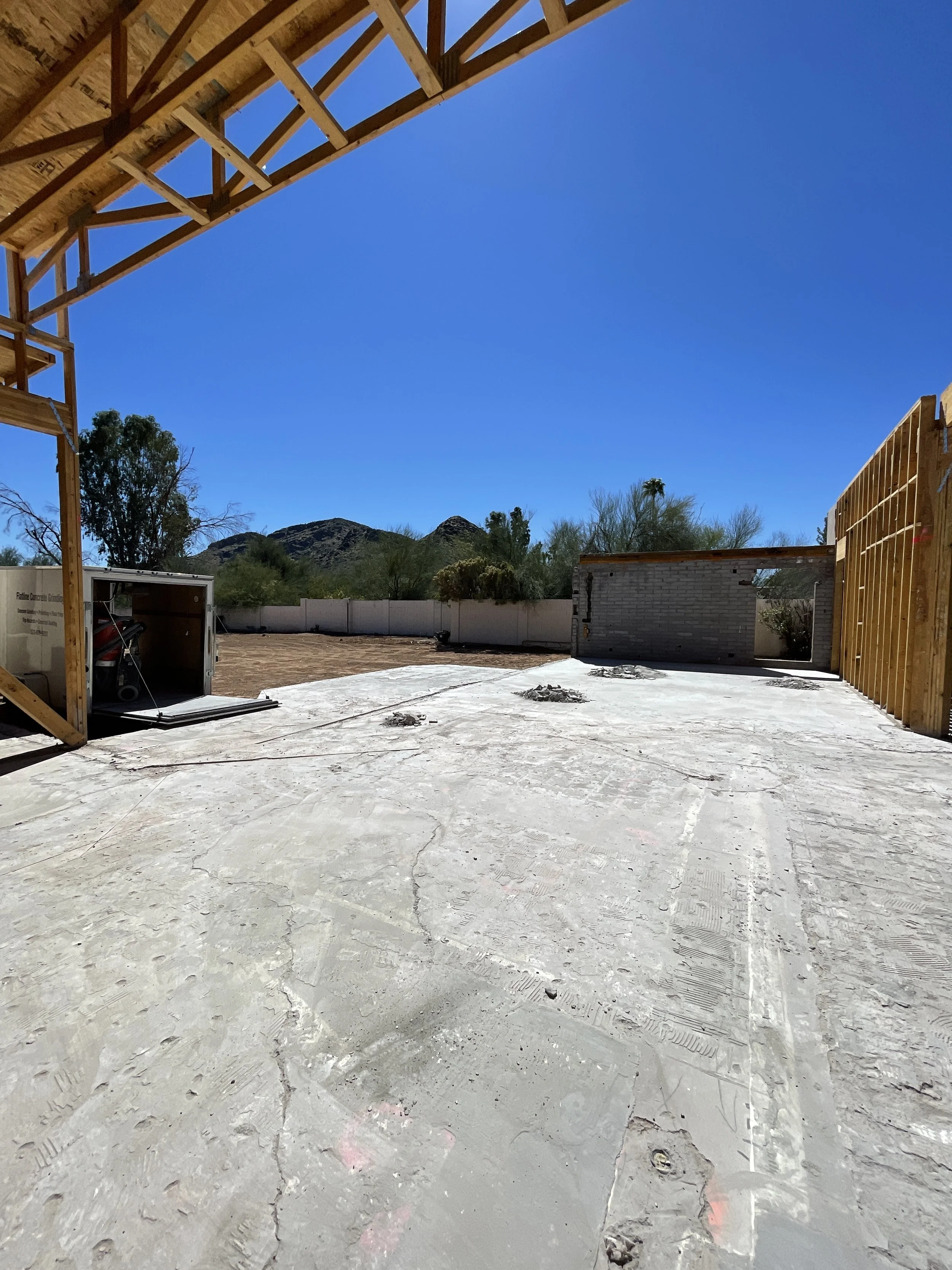 View of a construction site showing a concrete floor with cracks, wooden framing on the right, and a partially built wall on the right side. In the background, there are trees, mountains, and a blue sky.